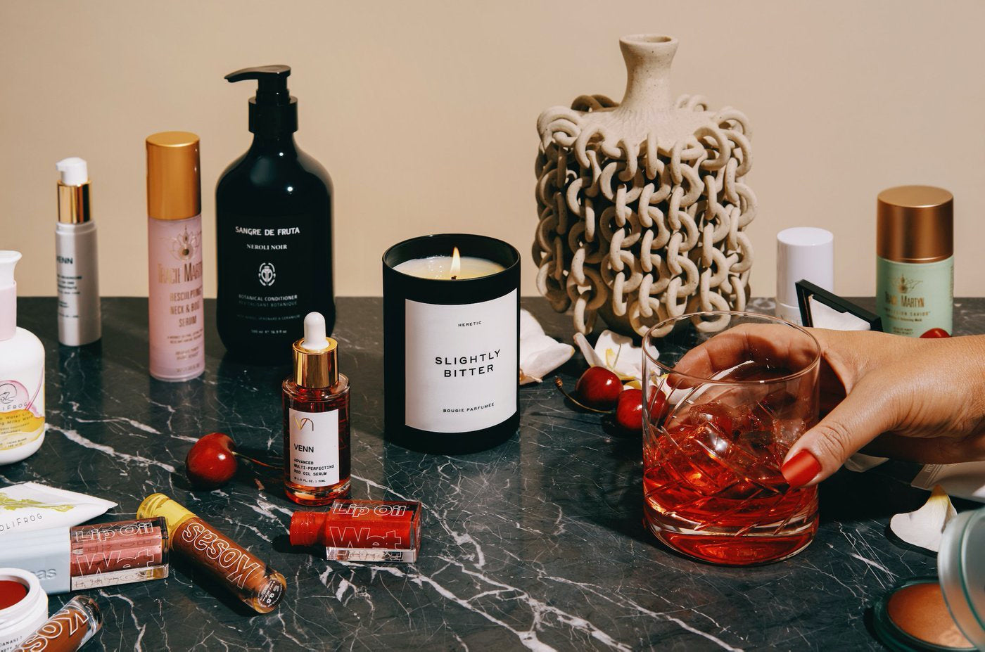 An array of cosmetics products on a table. A hand holding a drink enters the frame