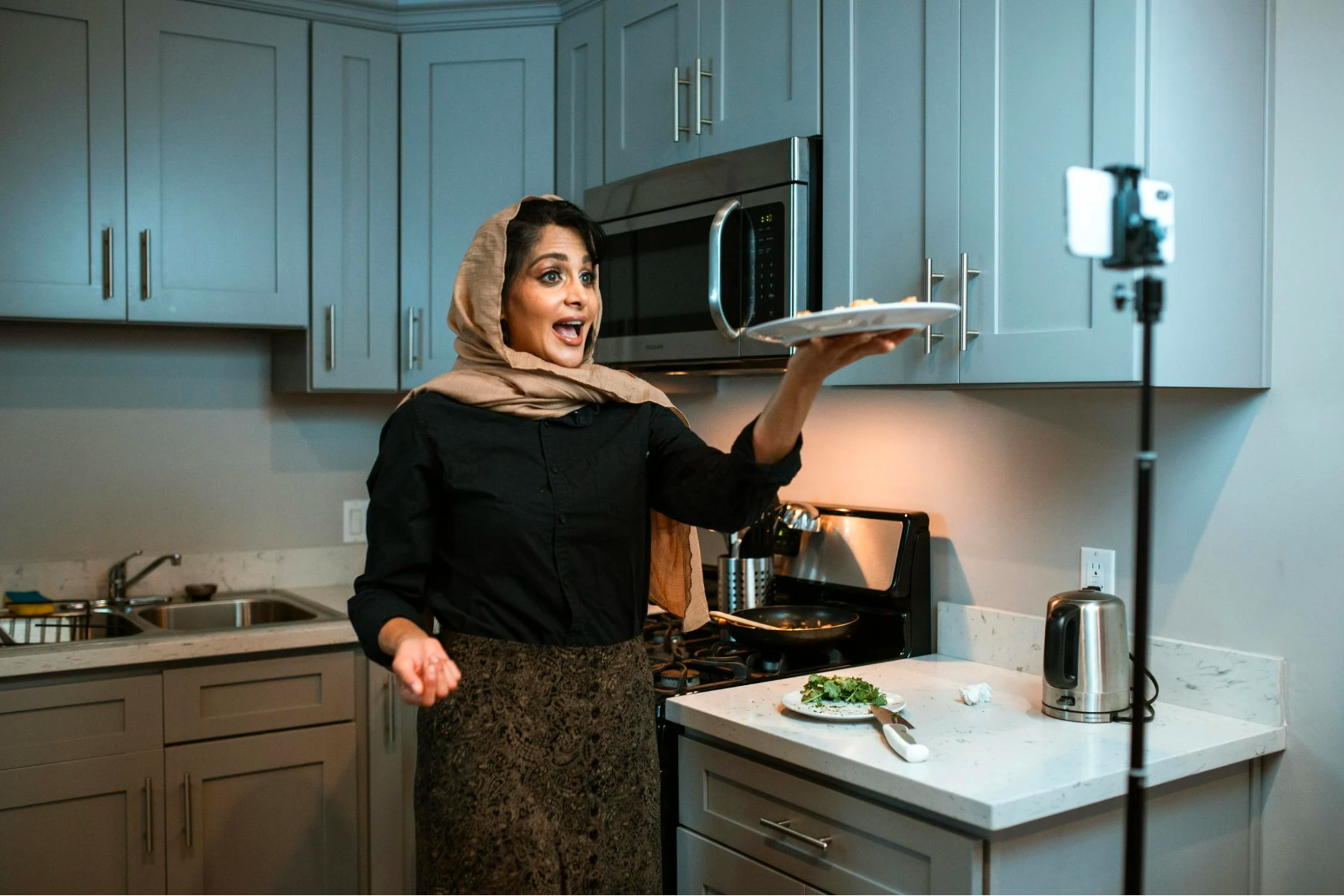 An influencer records content from a kitchen, holding a plate of food to a smartphone mounted on a stand.