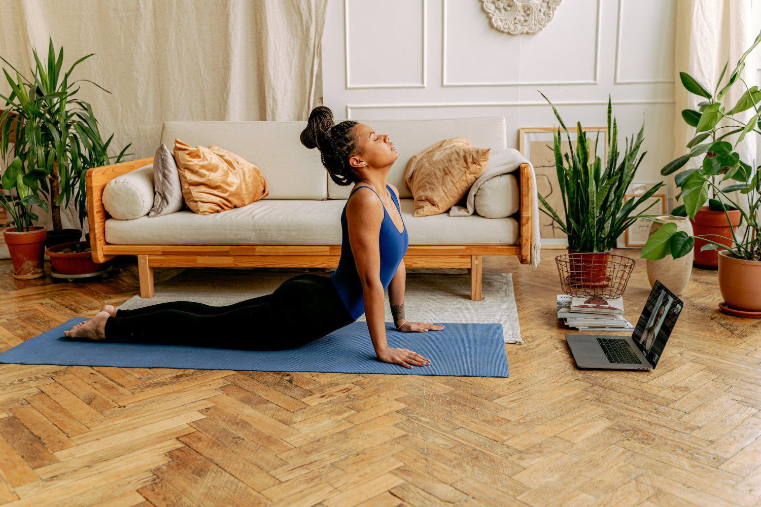 A person performs a yoga pose in her living room while watching a laptop