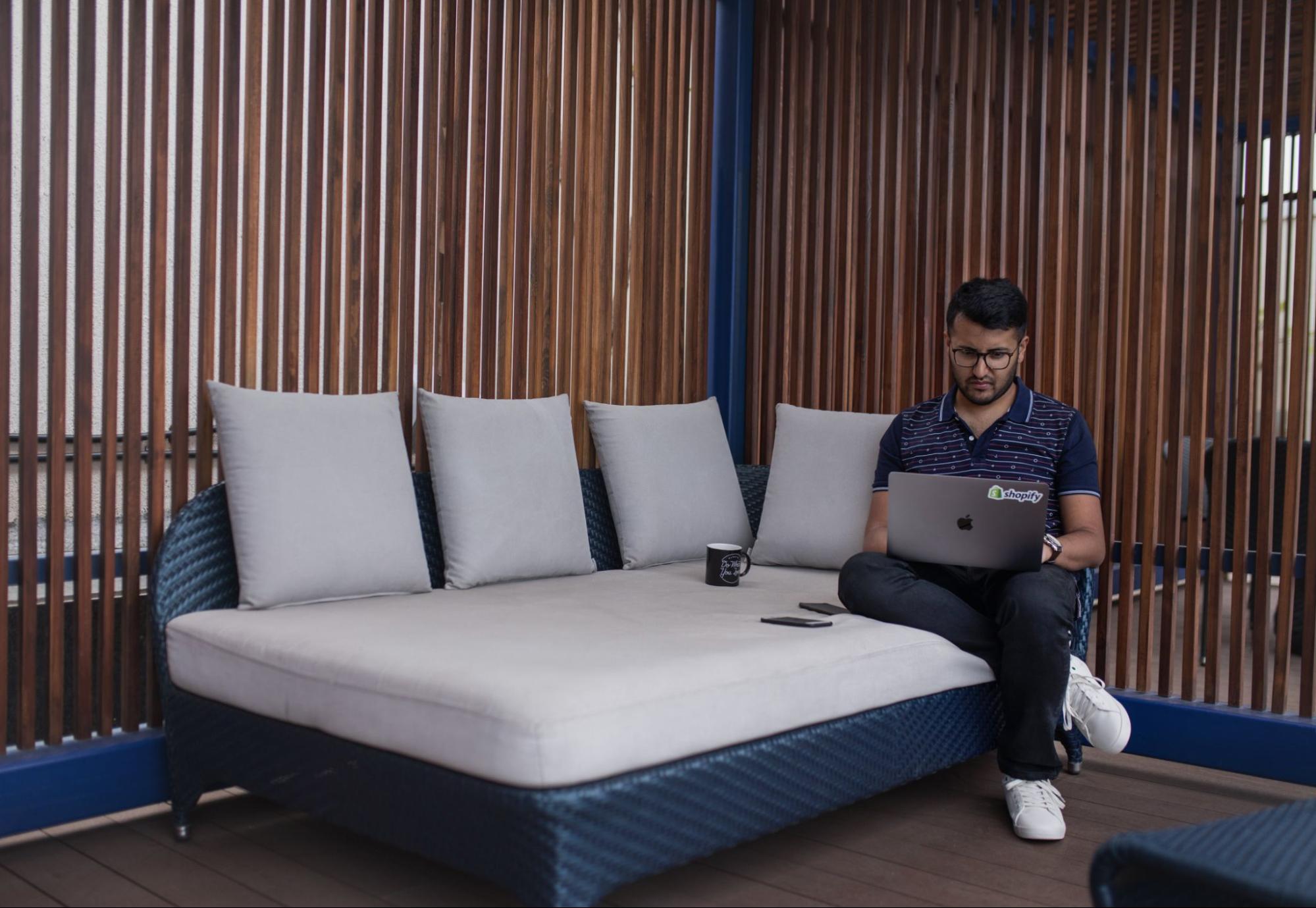 young man sits comfortably on a patio couch while working away on his laptop.