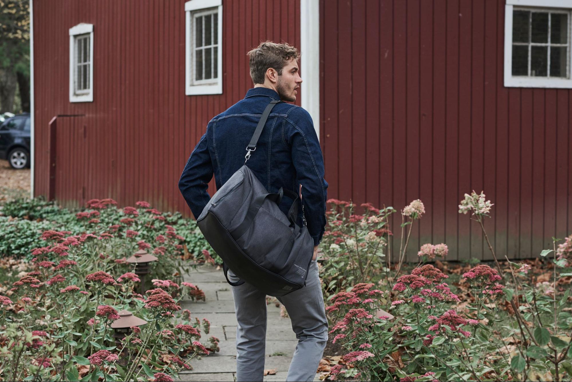 Lifestyle image of a man in a garden wearing a denim jacket with a duffel bag on his shoulder.