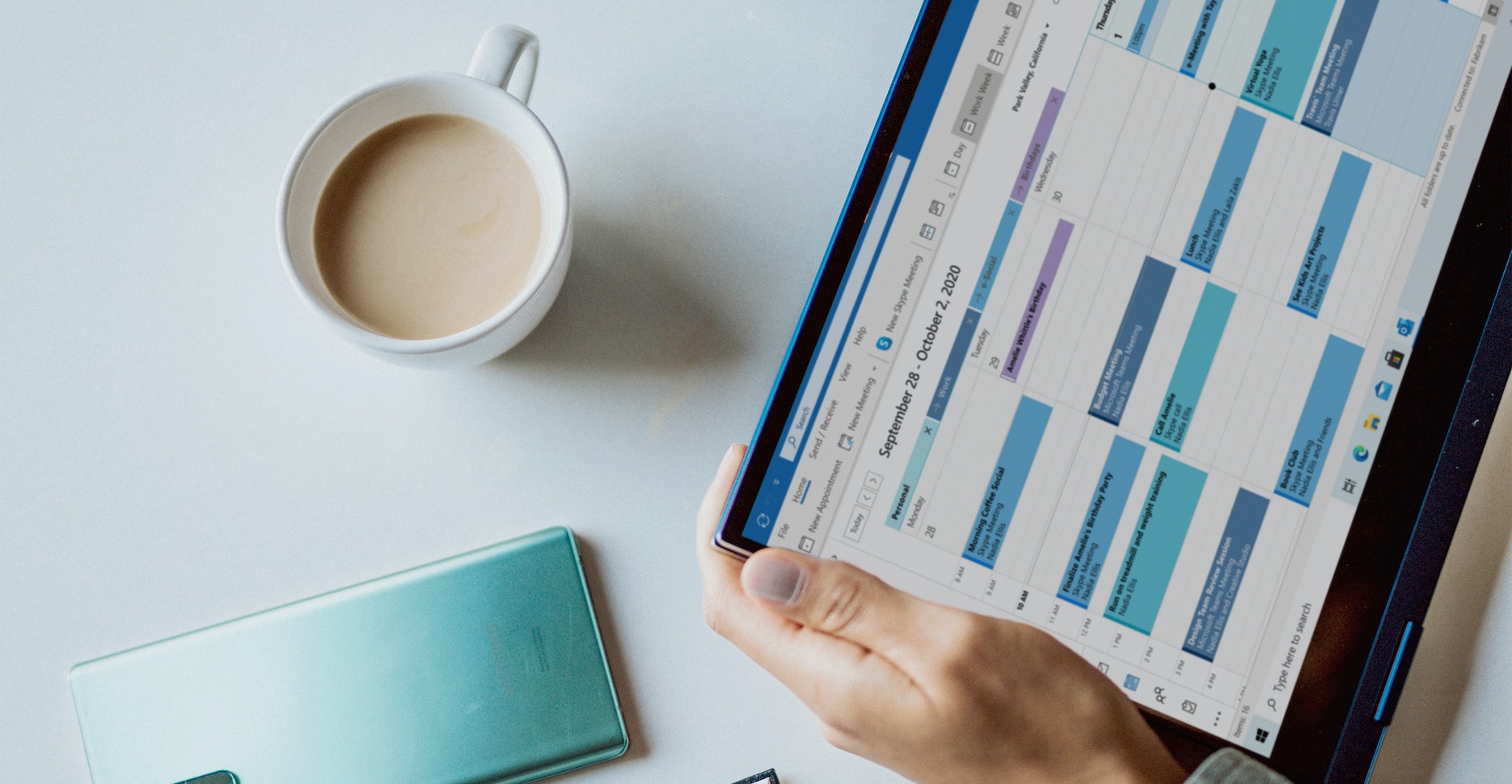 A person holds an ipad that shows a calendar with various appointments next to a cup of coffee