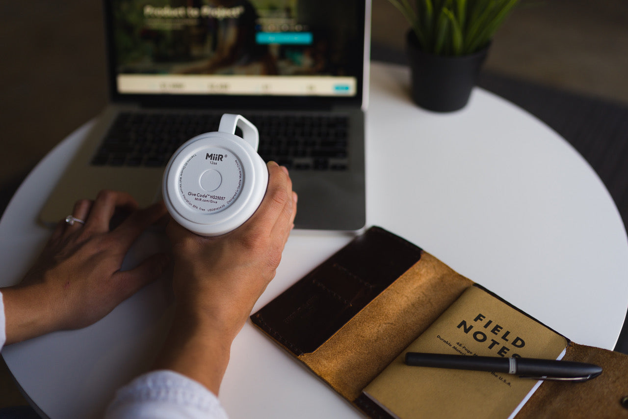 A MiiR mug being held with the top facing the camera, with a laptop in the background