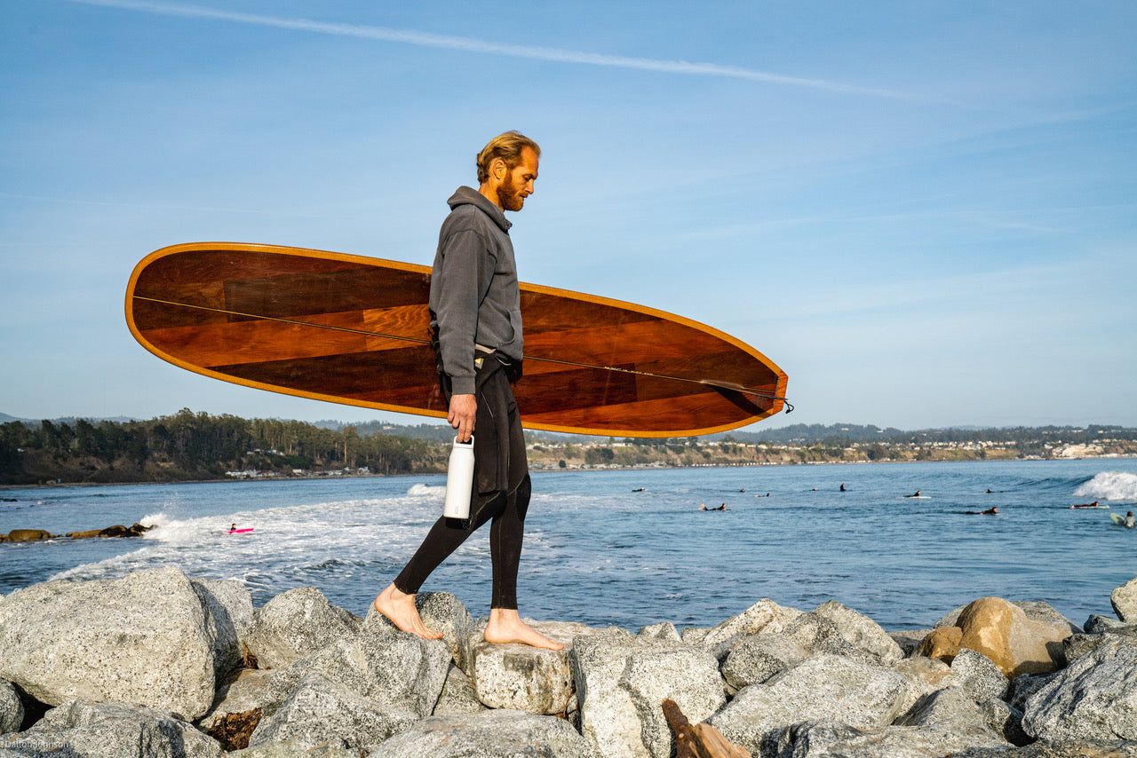 A surfer walking on a rocky pier holding a surfboard and a white MiiR drink thermos