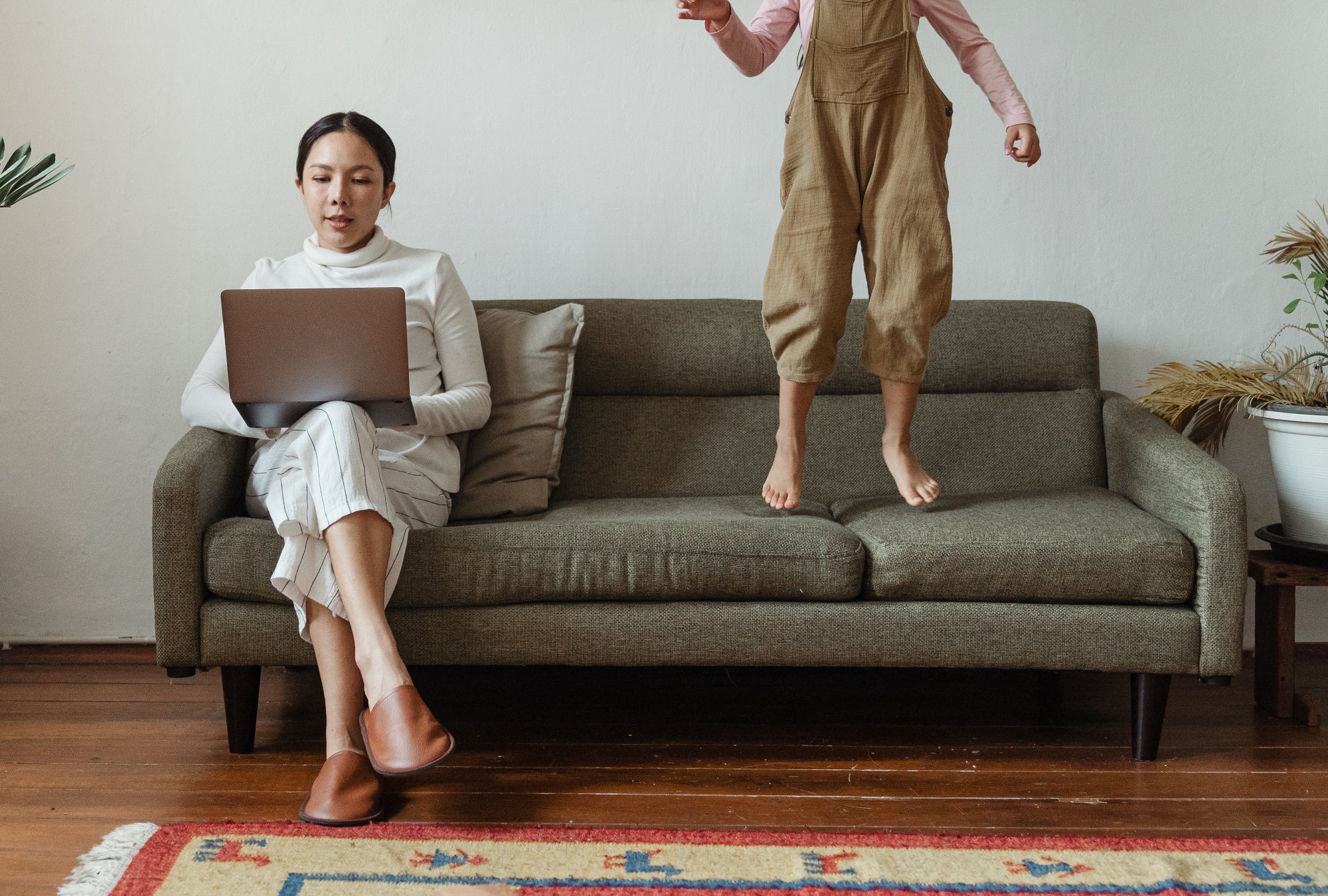 A mother works on a laptop while her child jumps on the couch beside her