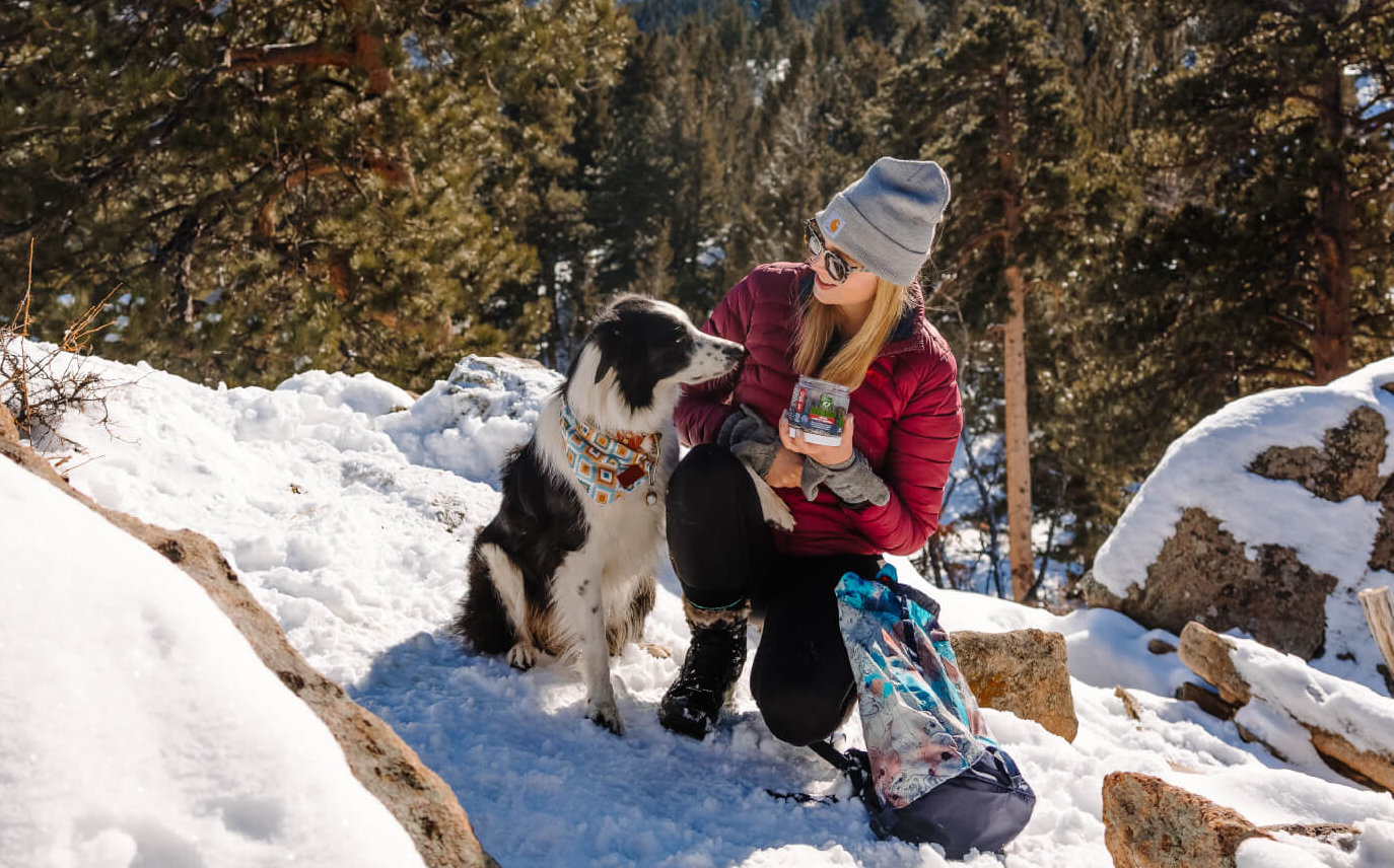 In a snowy mountain scene, a dog owner prepares a snack for their eagerly awaiting pup.
