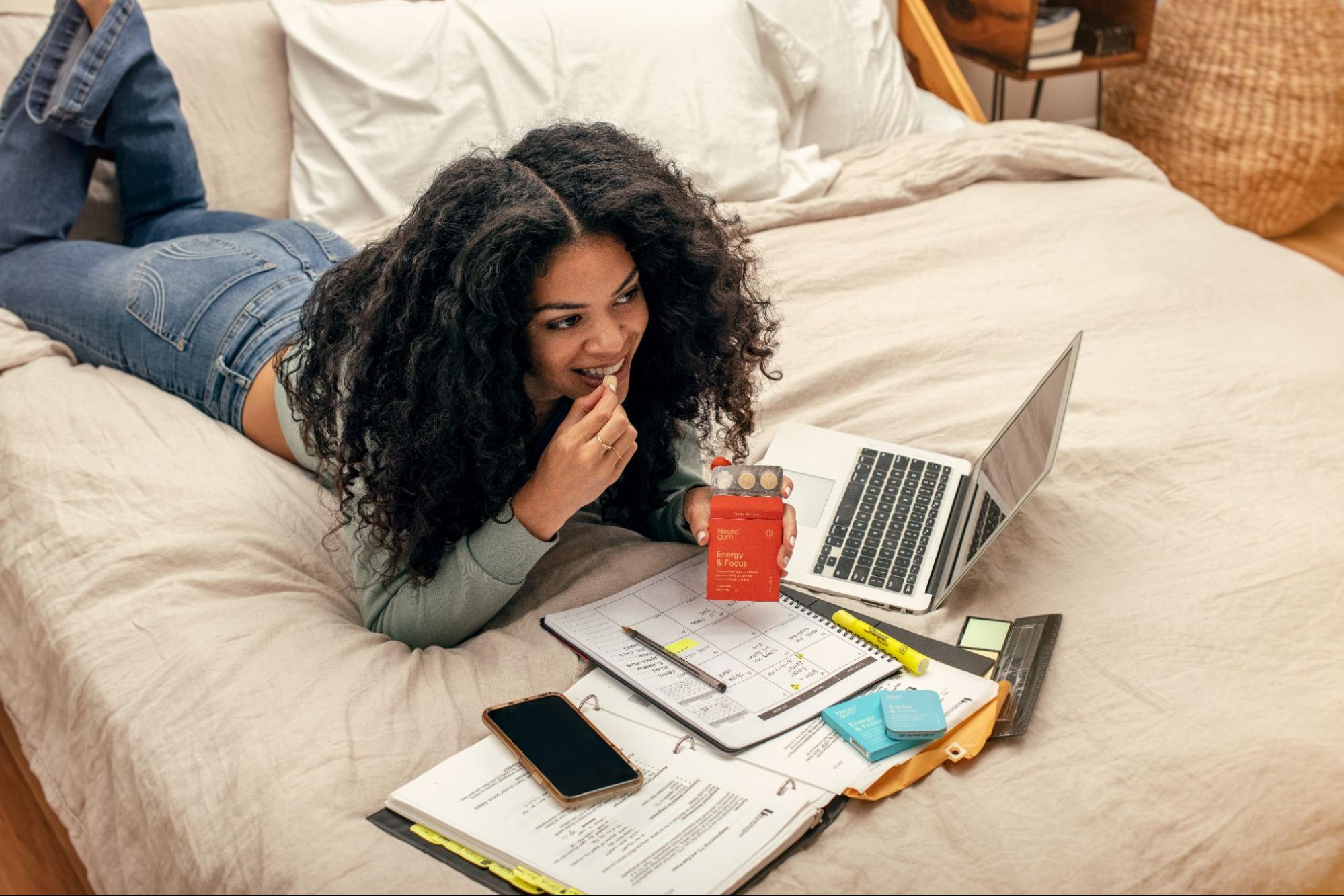 A model studying in bed while holding up a pack of Neuro gums. 