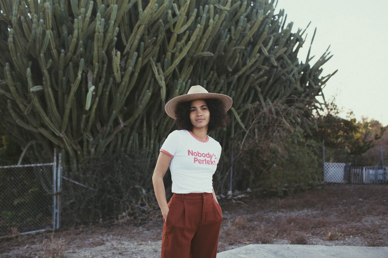 Een vrouw draagt een hoed en een T-shirt met de tekst "Nobody's Perfekt" en staat voor een achtergrond met bomen