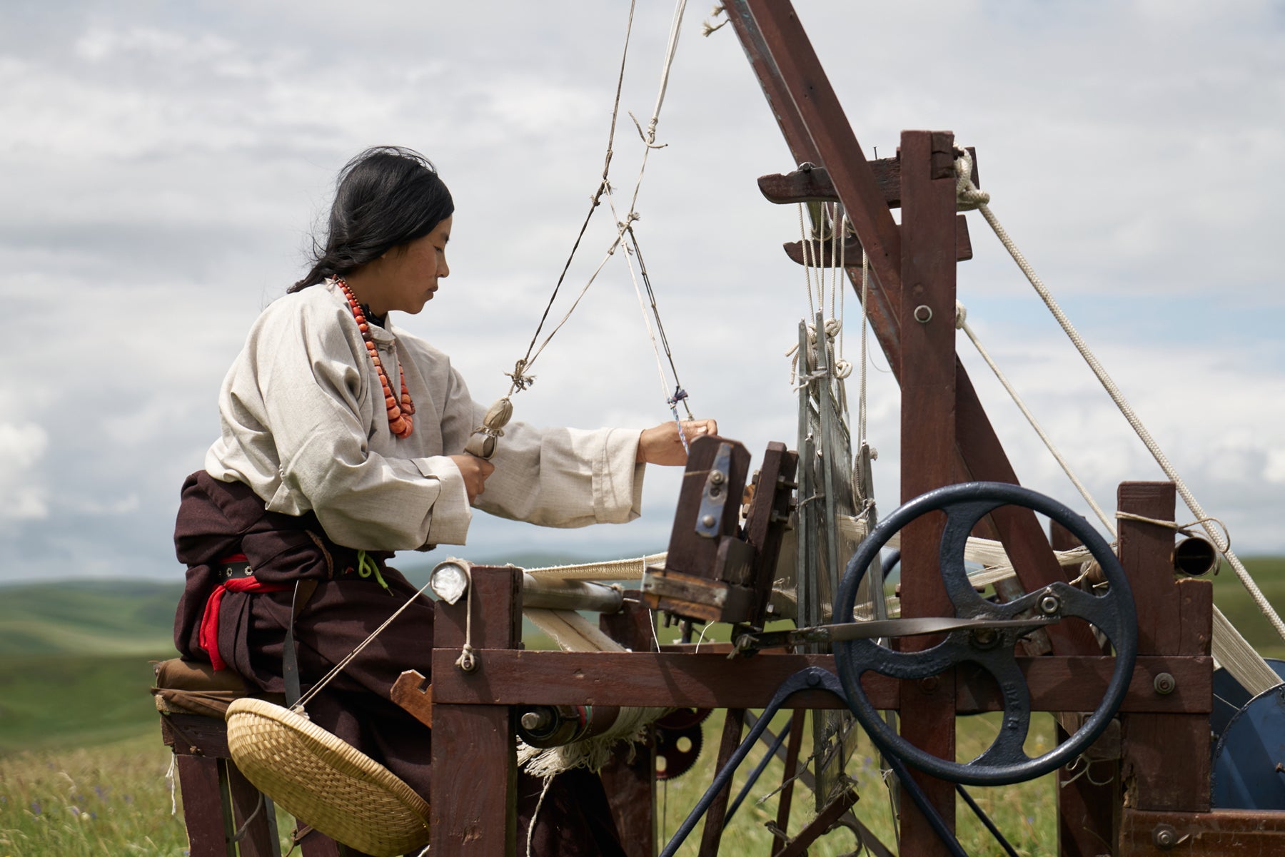 An artisan, in beige shirt and brown pants at an outdoor weaving machine, works on a piece of textile. 