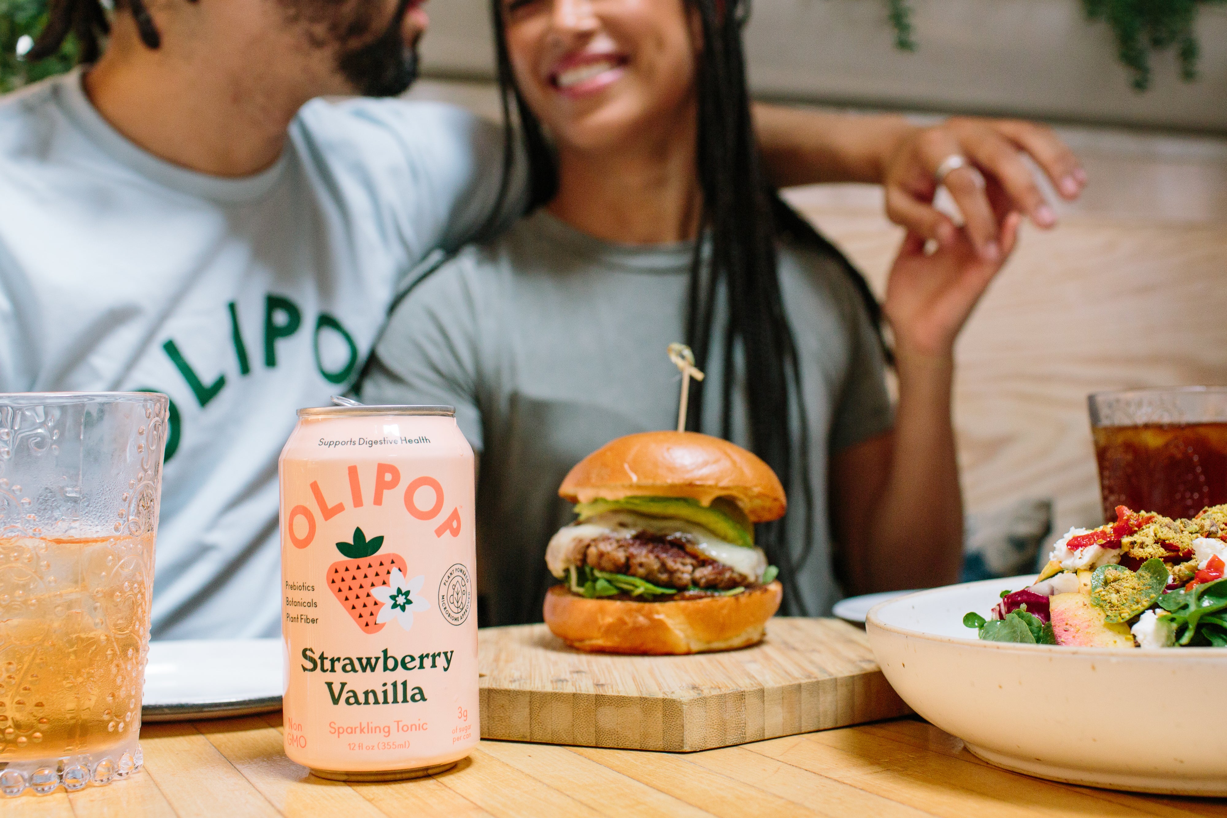 A couple embracing while sitting at a restaurant with a burger and a can of Olipop in front of them. 