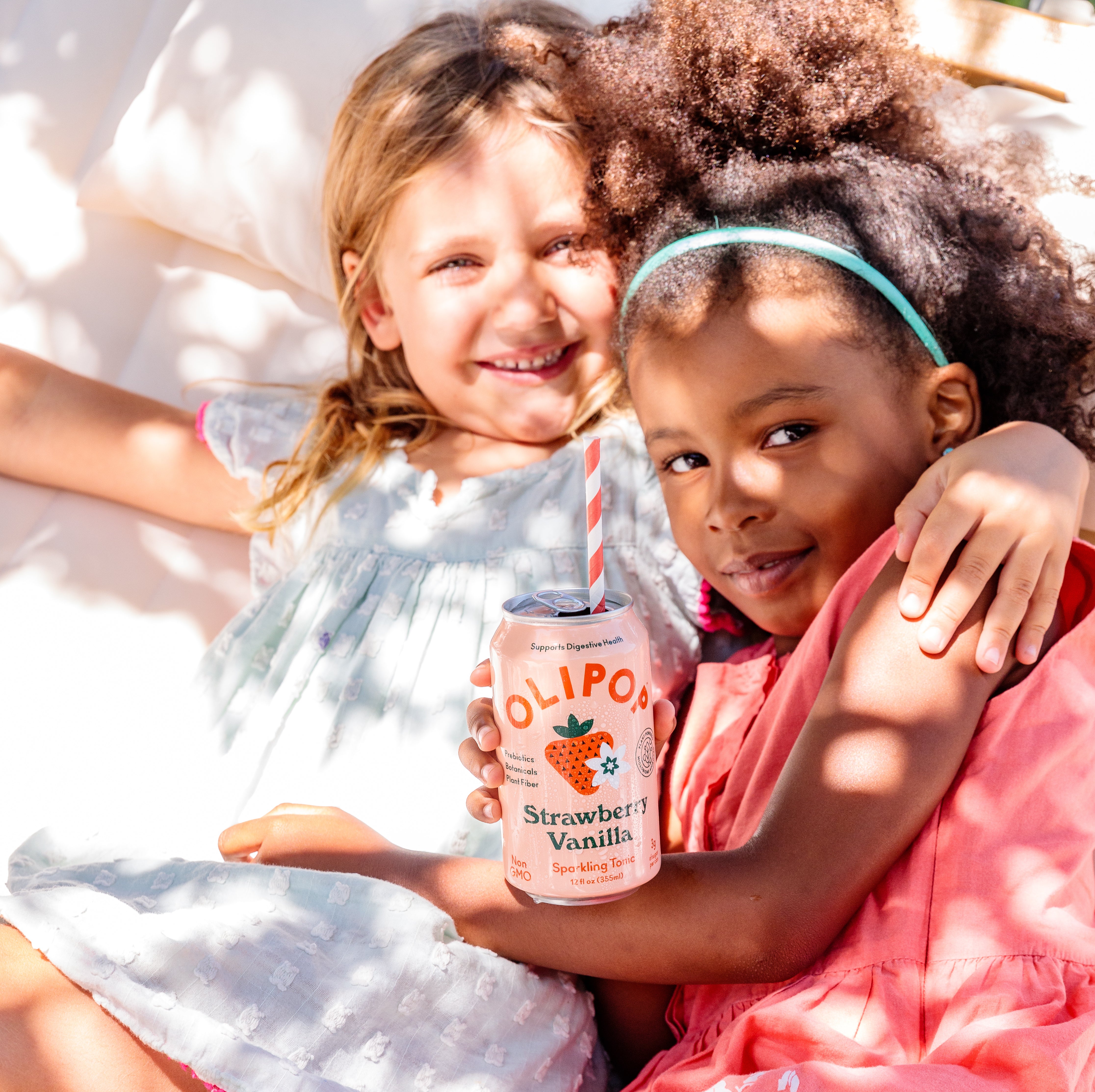 Two young girls lying on a picnic blanket embracing while drinking a can of Olipop.