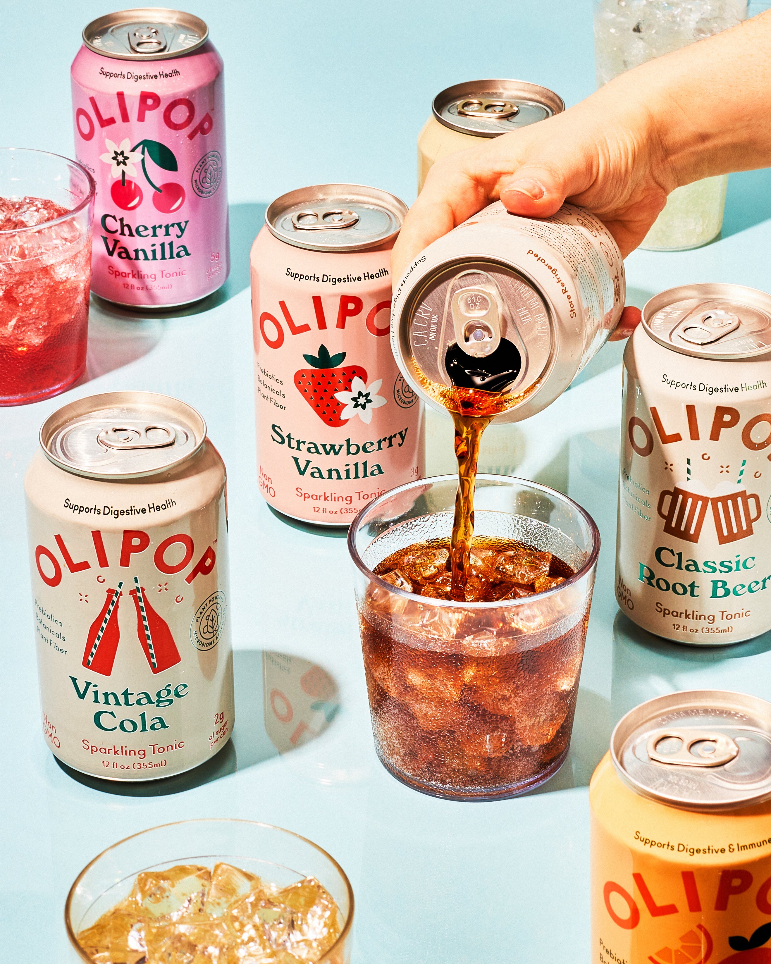 A hand pouring a drink into a glass that is surrounded by other cans/ flavours of Olipop