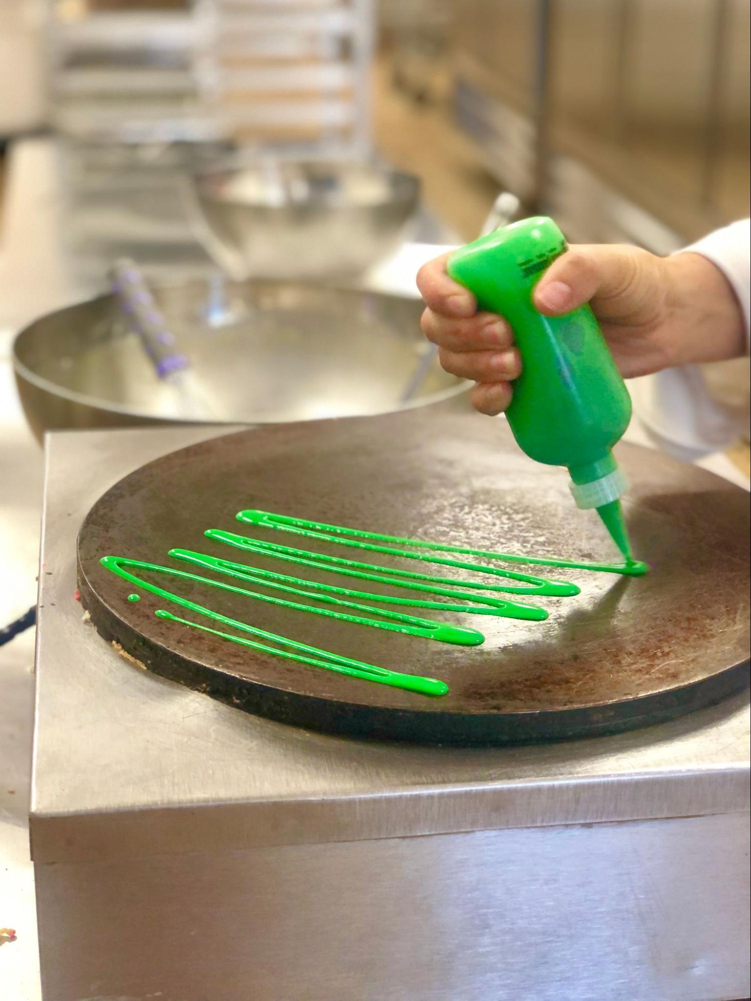 A hand squeezing a pastry bottle filled with green batter onto a crepes pan.