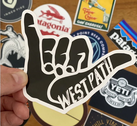 Hand holding a West Path sticker in the foreground with patagonia and yeti stickers in background.