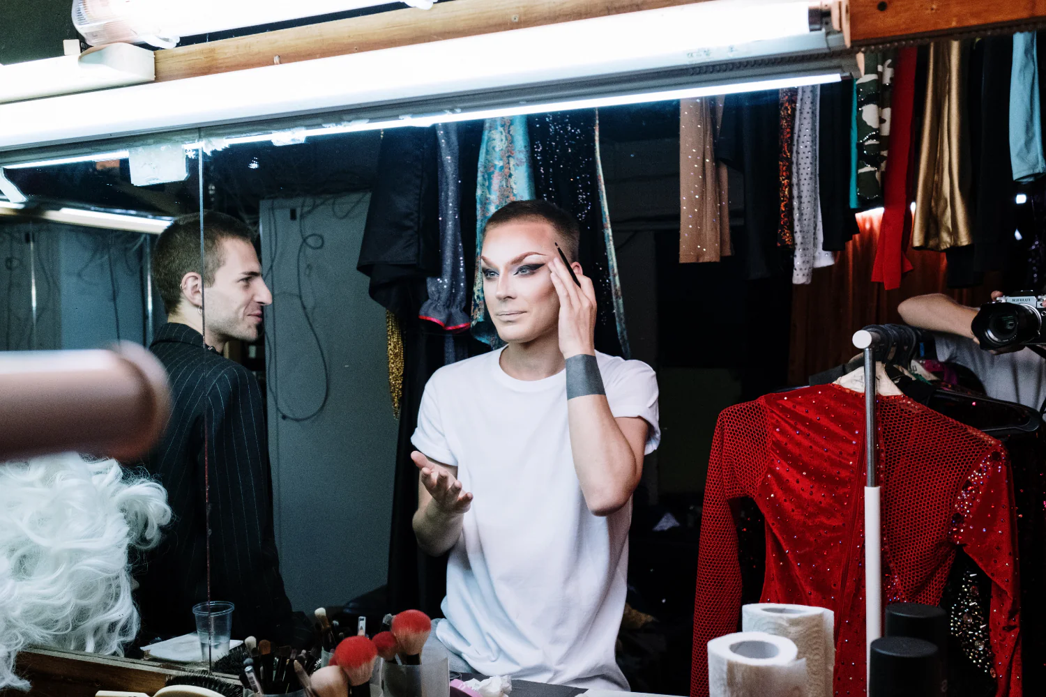 A person applies drag makeup in front of a mirror in a green room