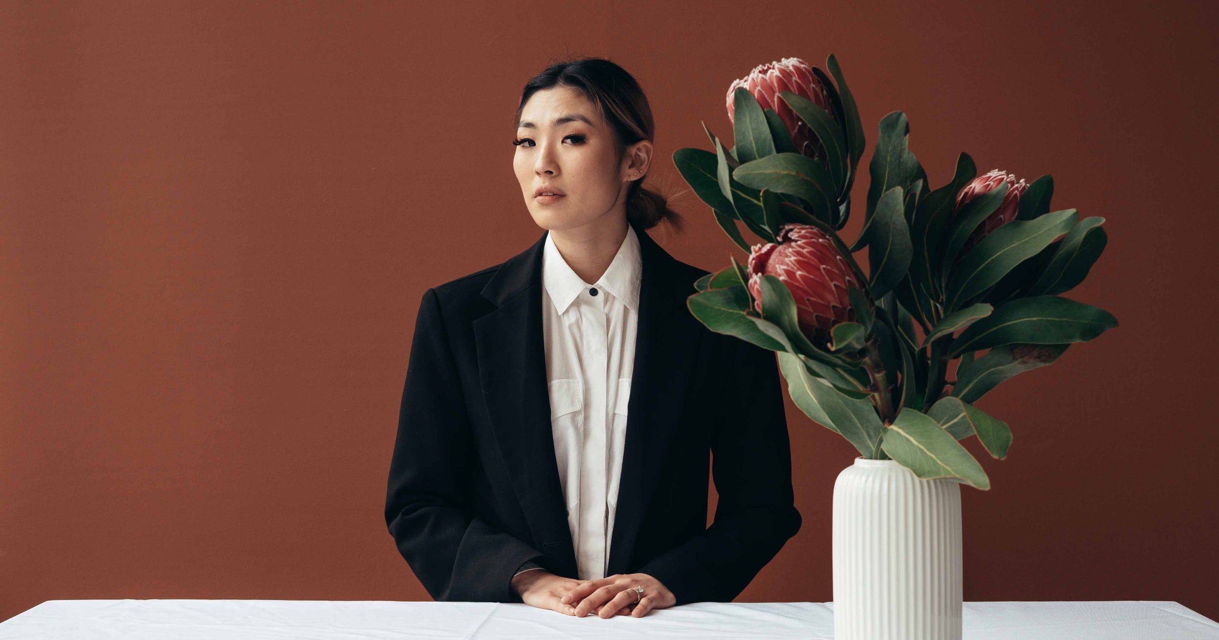 Woman wearing a blazer sits behind a table with a large vase of flowers perched on it