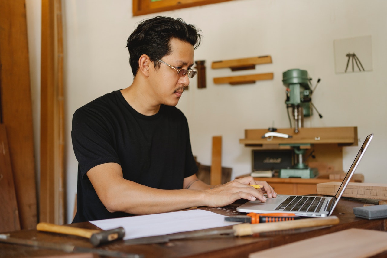 A man wearing glasses works at a laptop on a bench in a workshop