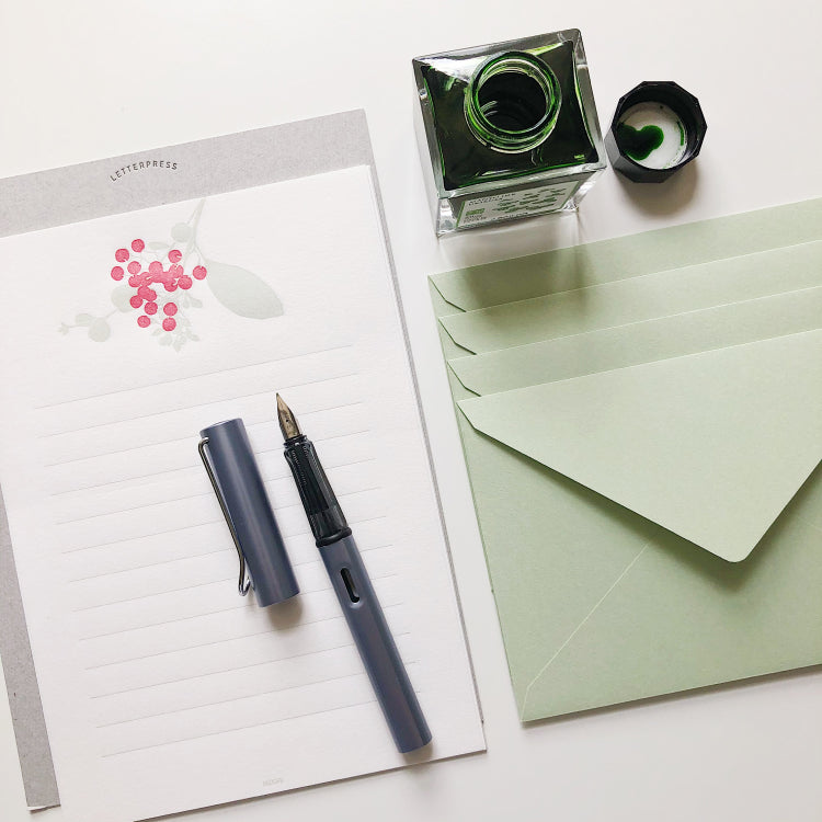 A fountain pen rests on a notepad. A green inkwell is at the top of the image with green envelopes to the right.