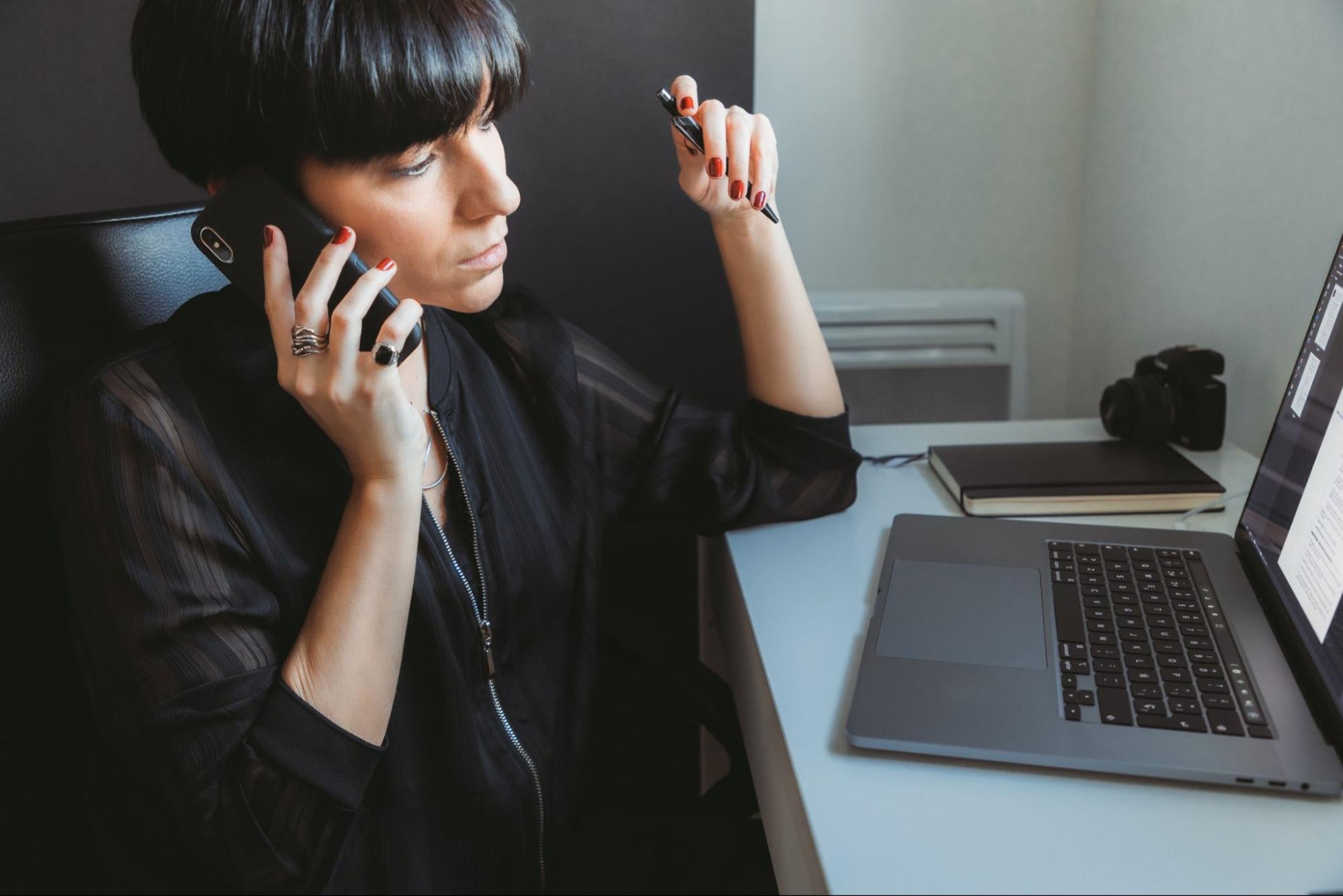 A woman sits at her desk looking at a computer screen as she holds a phone to hear ear and a pen up in the other hand.