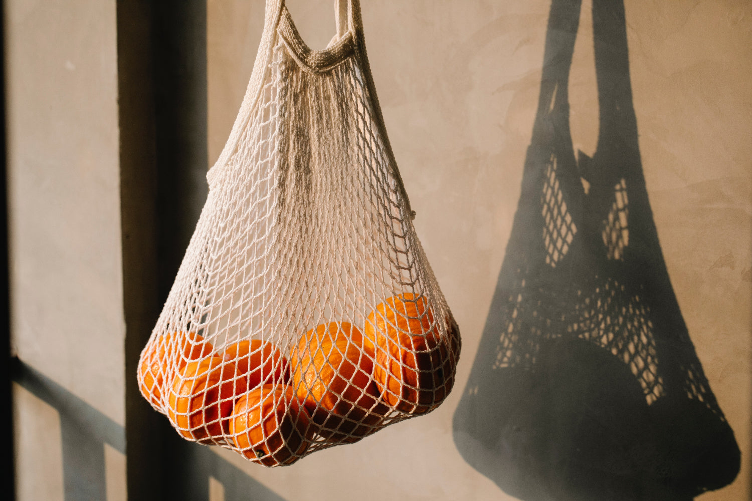 A mesh bag full of oranges casts a shadow on a beige wall