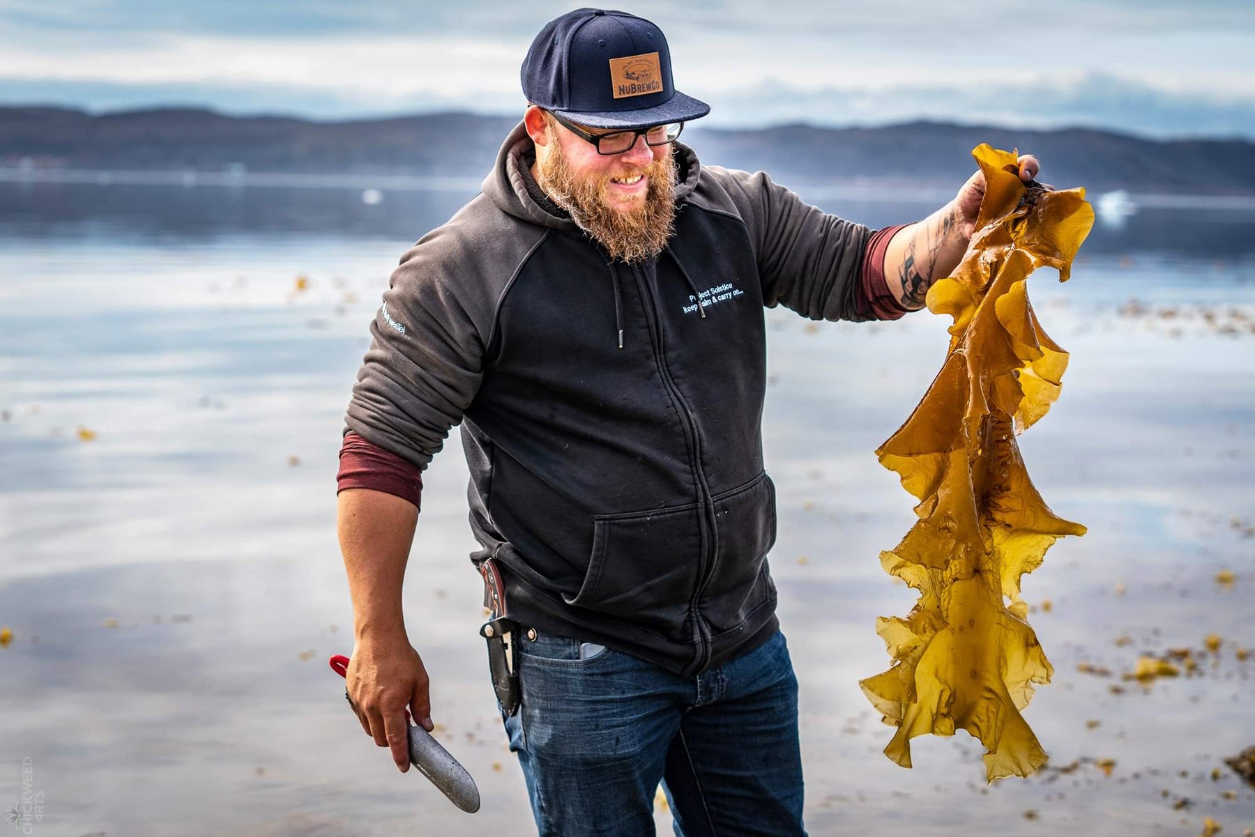 Photograph of Justin Clarke of UasaU Soap, smiling at the large piece of seaweed that he's recently foraged in Nunavut, where UasaU soap is produced. 