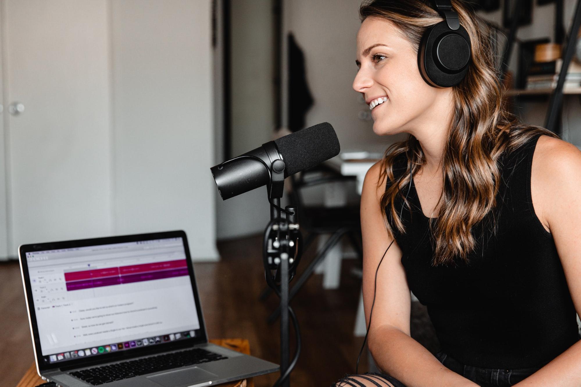 A woman wearing a black tank top sitting in front of a microphone and open laptop recording a podcast