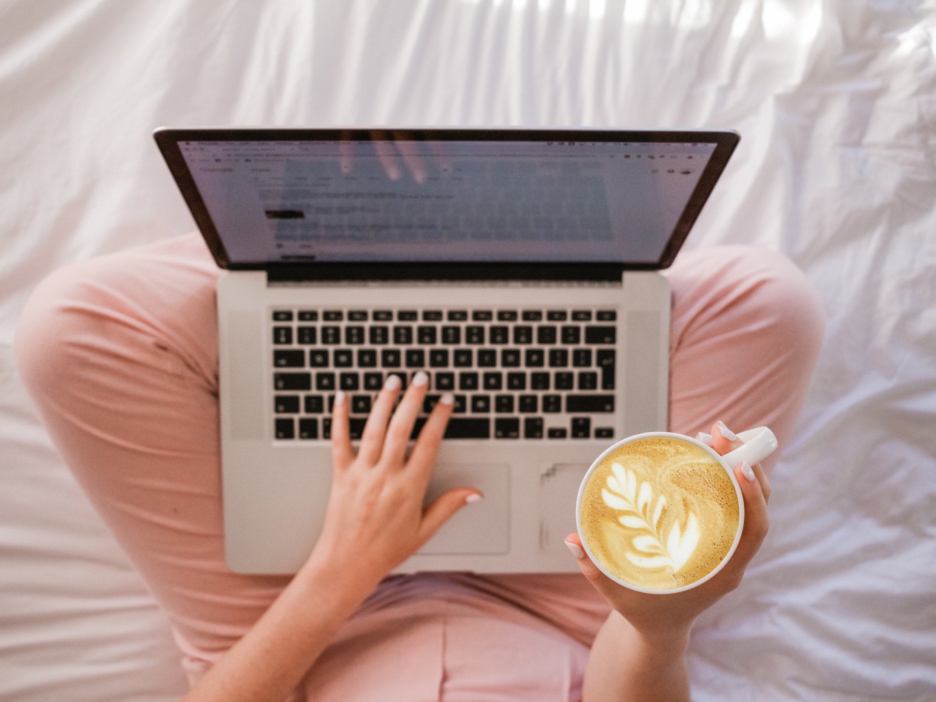 A person sits cross legged on a bed with their laptop on their lap holding a latte
