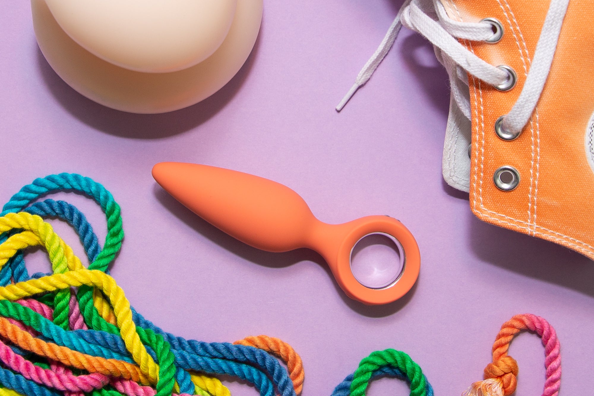 Das Foto zeig einen orangenen Vibrator, ein Regenbogenfarbenes Seil und einen orangenen Sneaker. Es dient zu Präsentation, um Sexspielzeug zu verkaufen