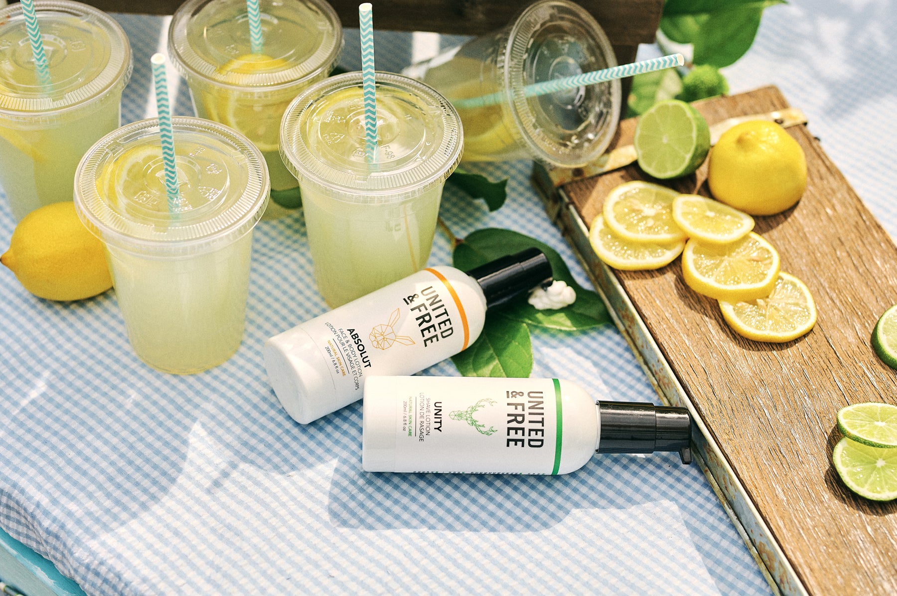 Two bottles of United & Free face and body lotion on a sunny tabletop surrounded by lemonade, lemons and limes.