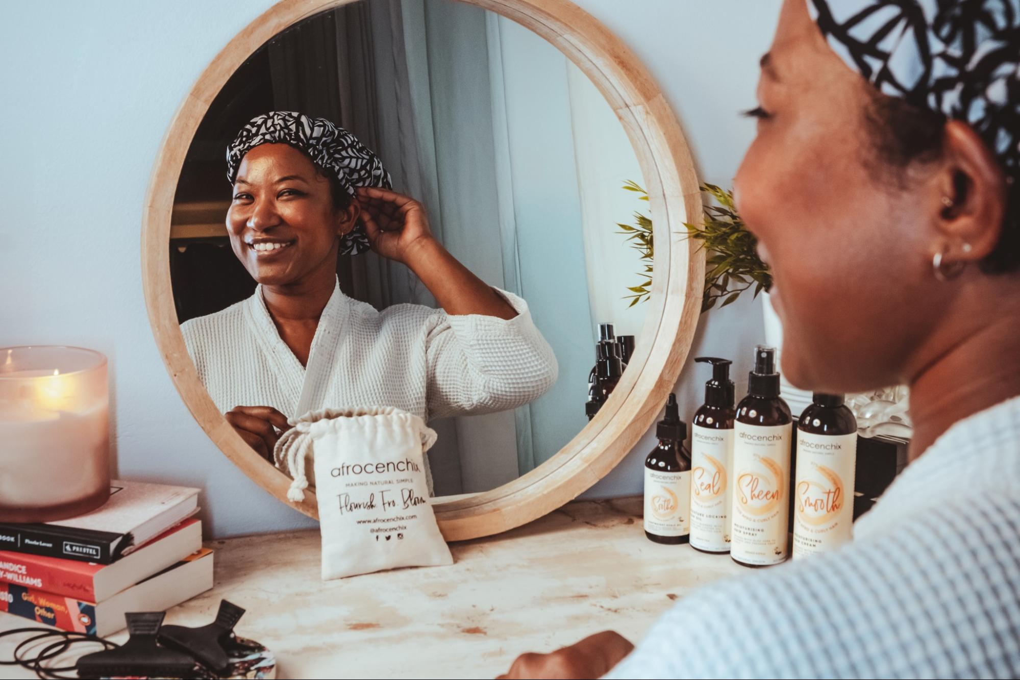 A model looks into the mirror while adjusting her bonnet from Afrocenchix, the table in front of her has products from Afrocenchix, books, and a candle. 