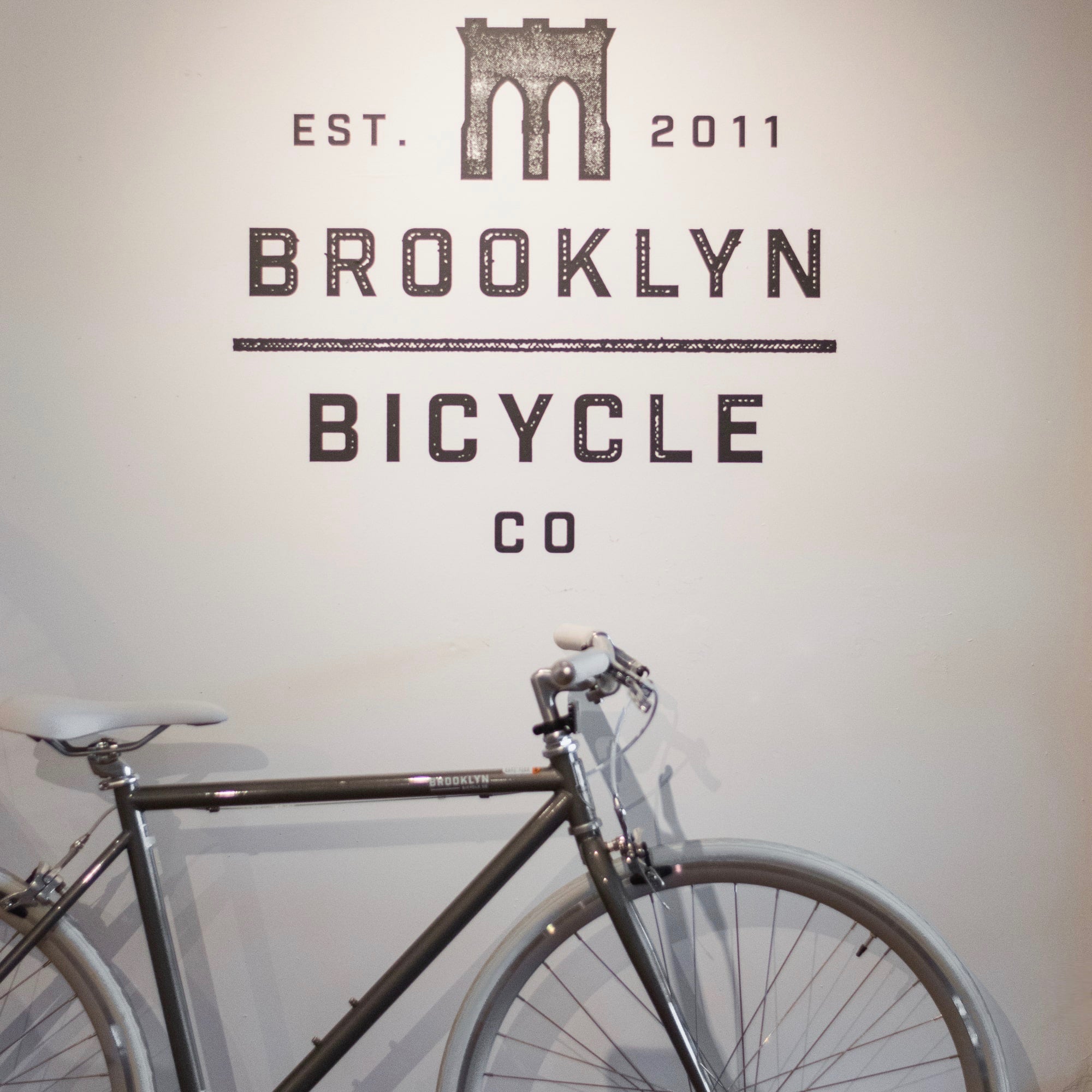 A grey bicycle by Brooklyn Bicycle Co. in their showroom against a wall with their logo. 