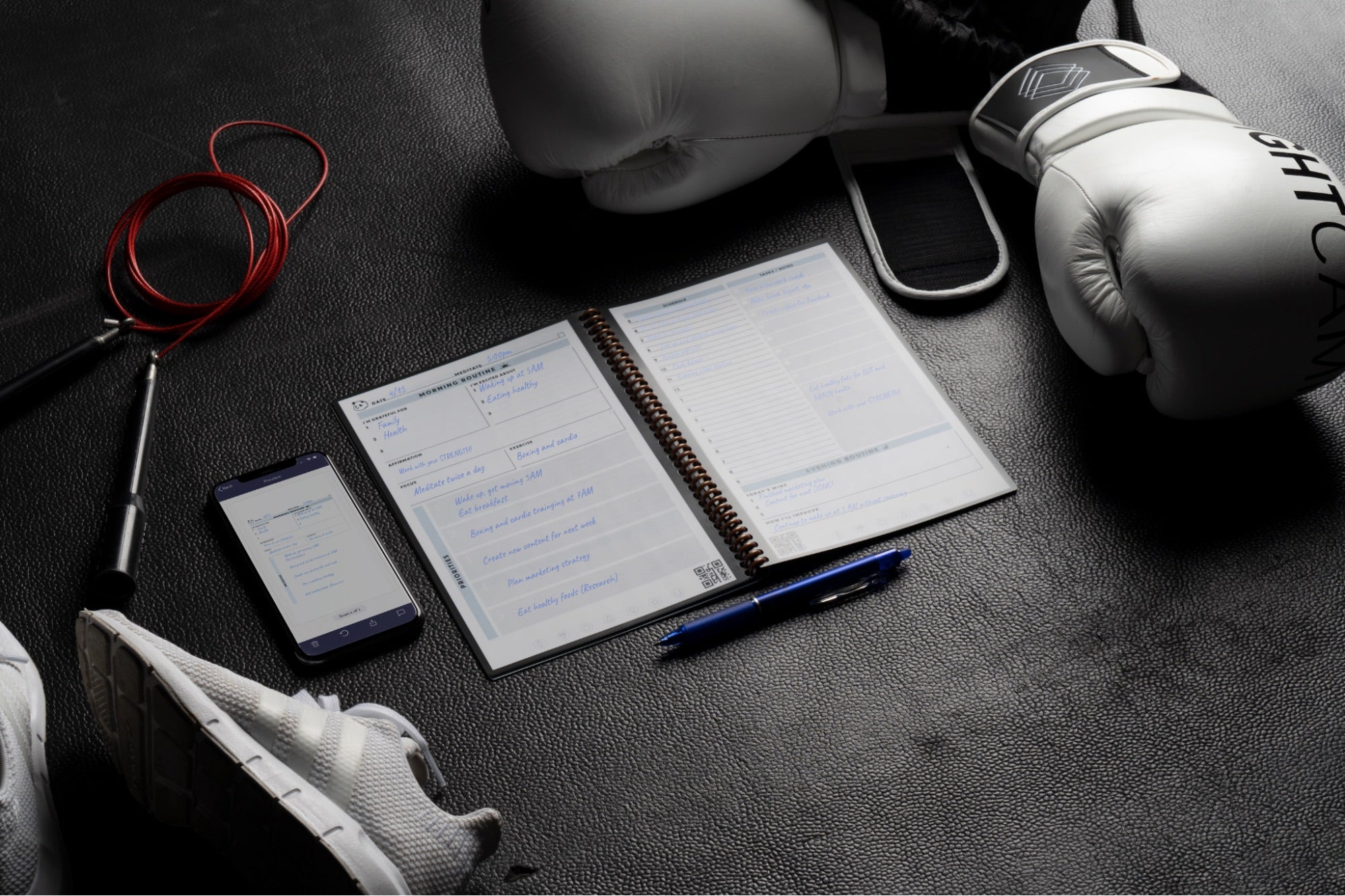 A Rocketbook notebook along with a smartphone displaying its app backdropped with workout gear like boxing gloves and running shoes.
