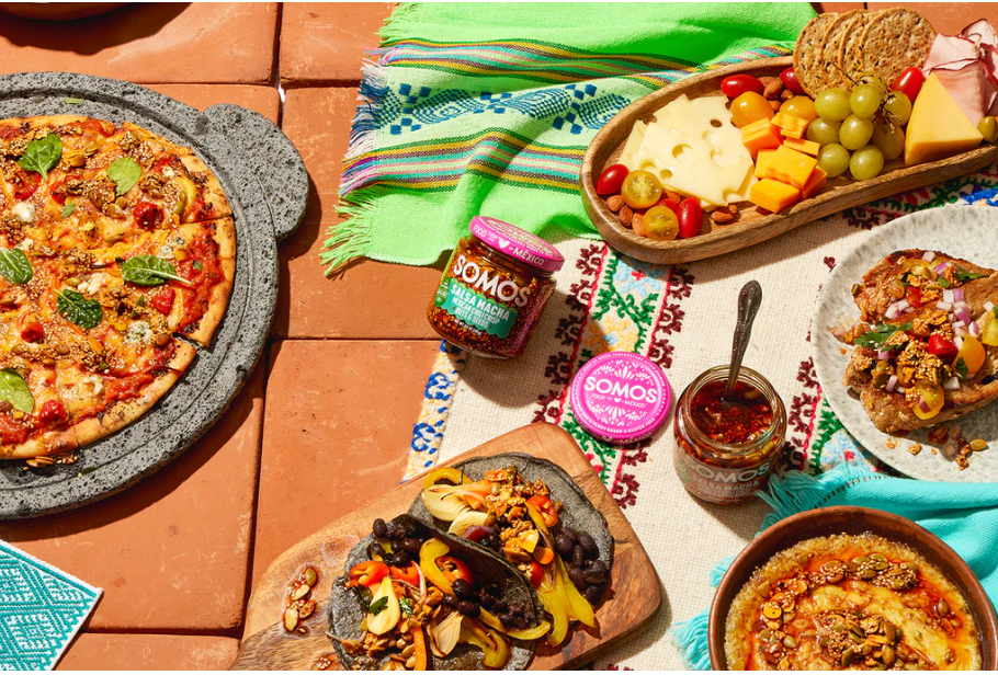 Food items laid out including a pizza, tacos, cheese and grapes, a tortilla, and jars of salsa.