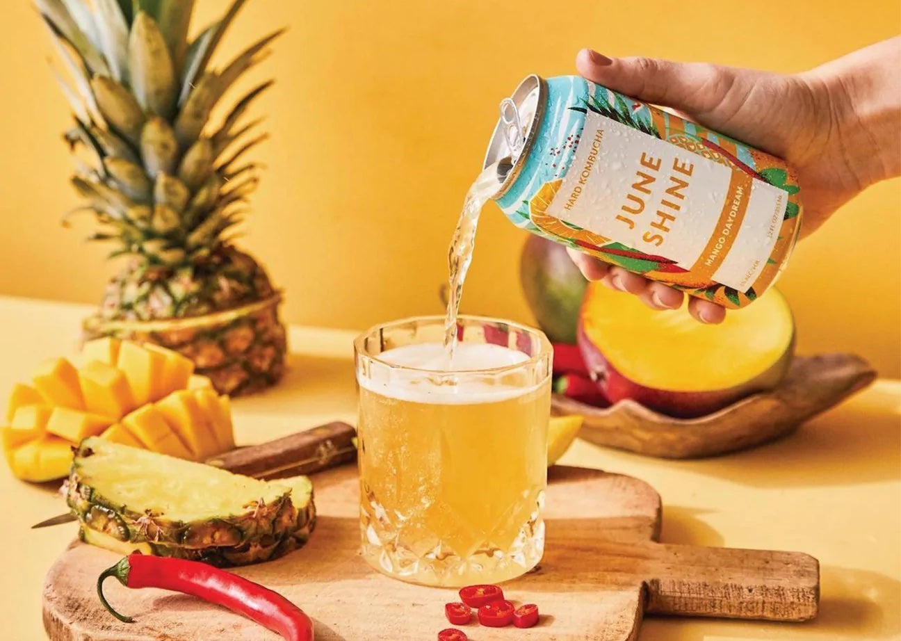 A hand pours a can of Juneshine into a glass. Fruits are arranged around the glass on a yellow background