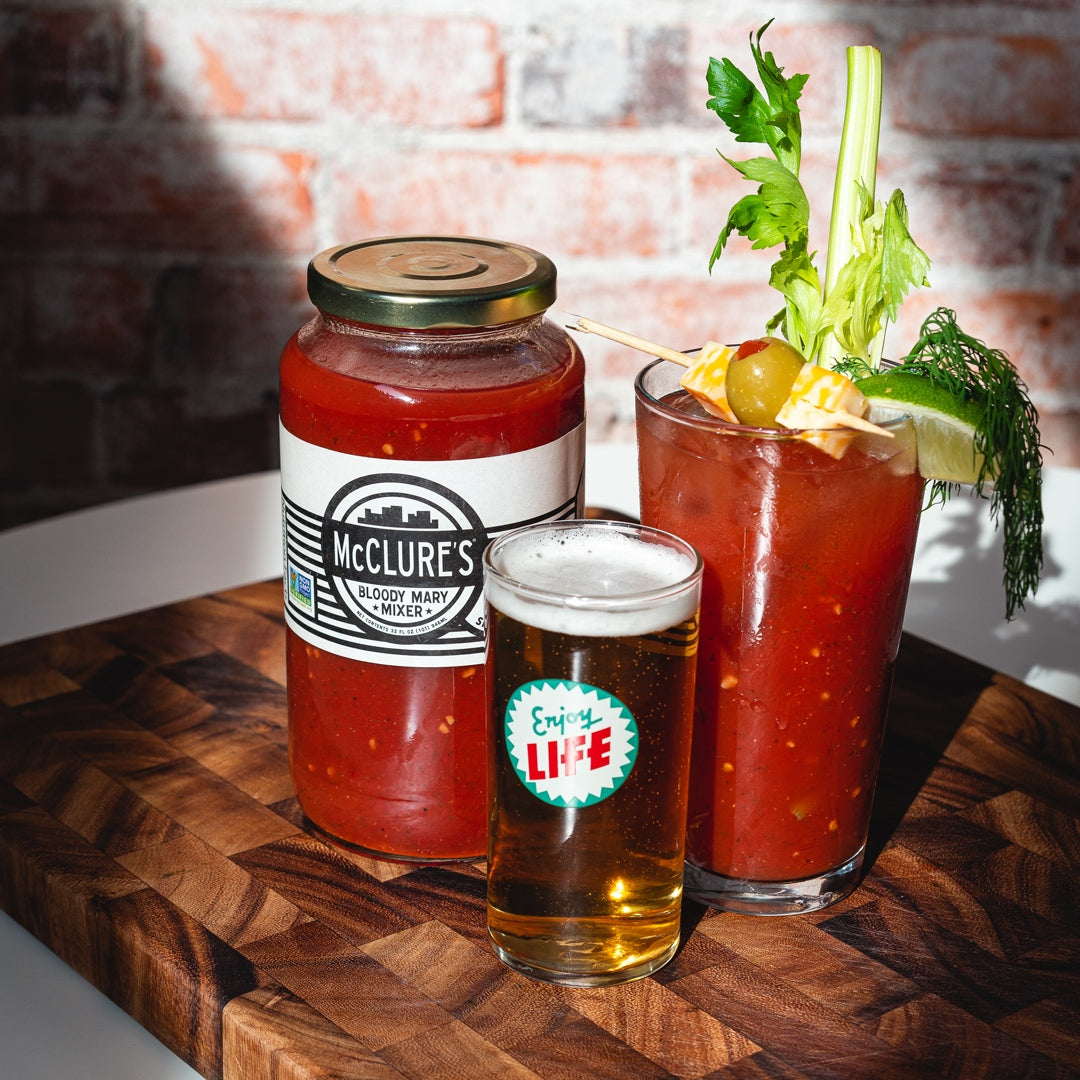 A jar of McClure's bloody mary mix, a mixed drink, and a beer are arranged on a wooden board