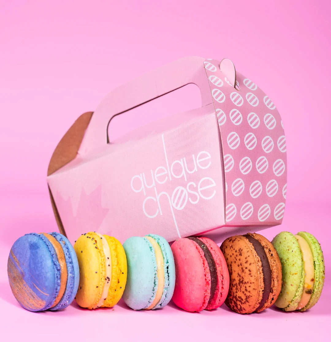 A pink box reads "Quele Chose." Colorful macarons are lined up in front of it