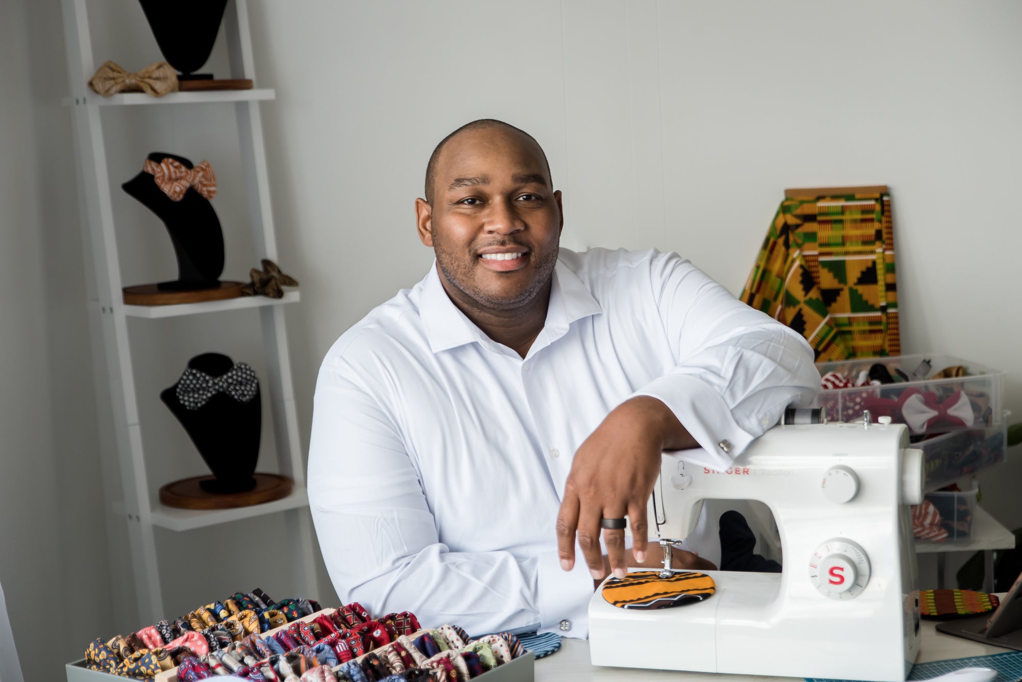 Founder Terrell Grayson leans on a sewing machine in a studio full of bow ties