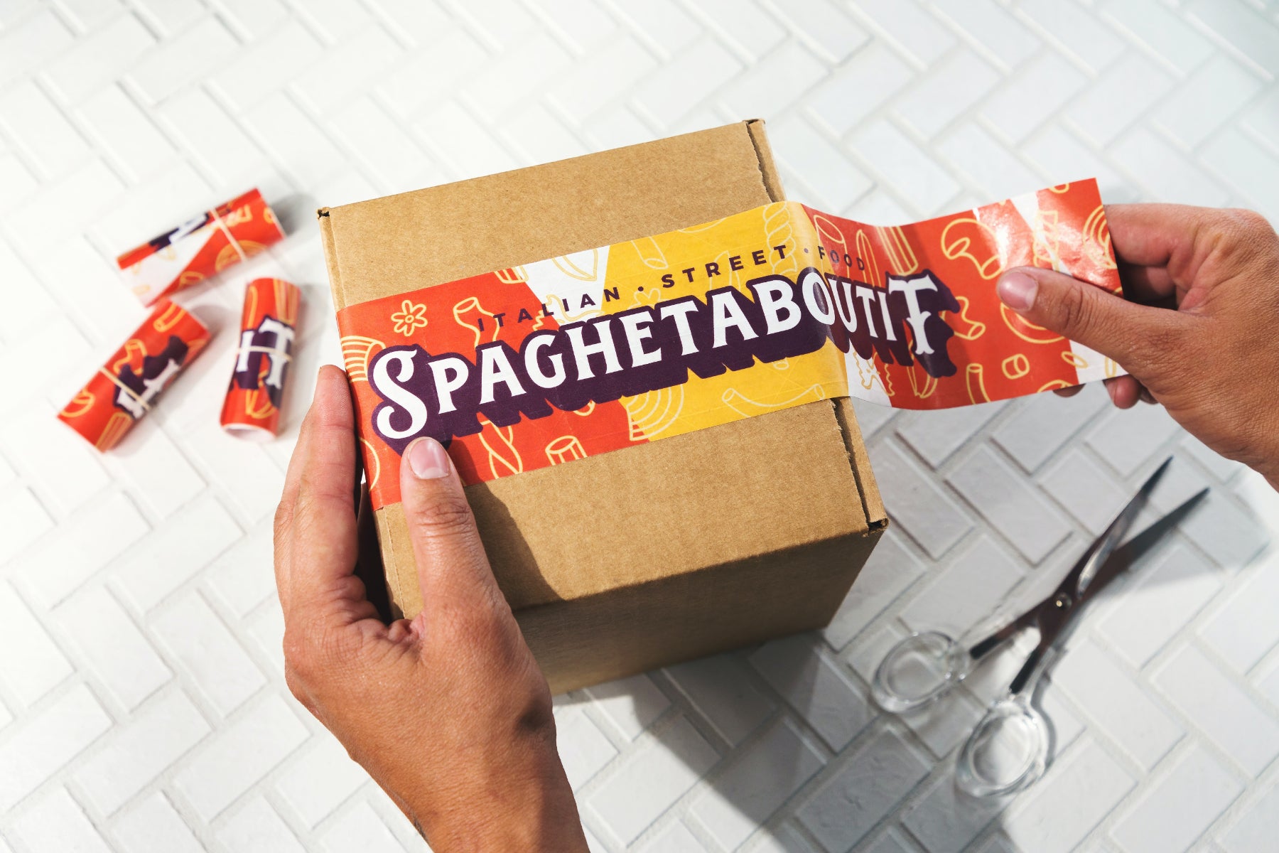 A person adds custom paper tape to a shipping box. The tape reads "Spaghetaboutit"