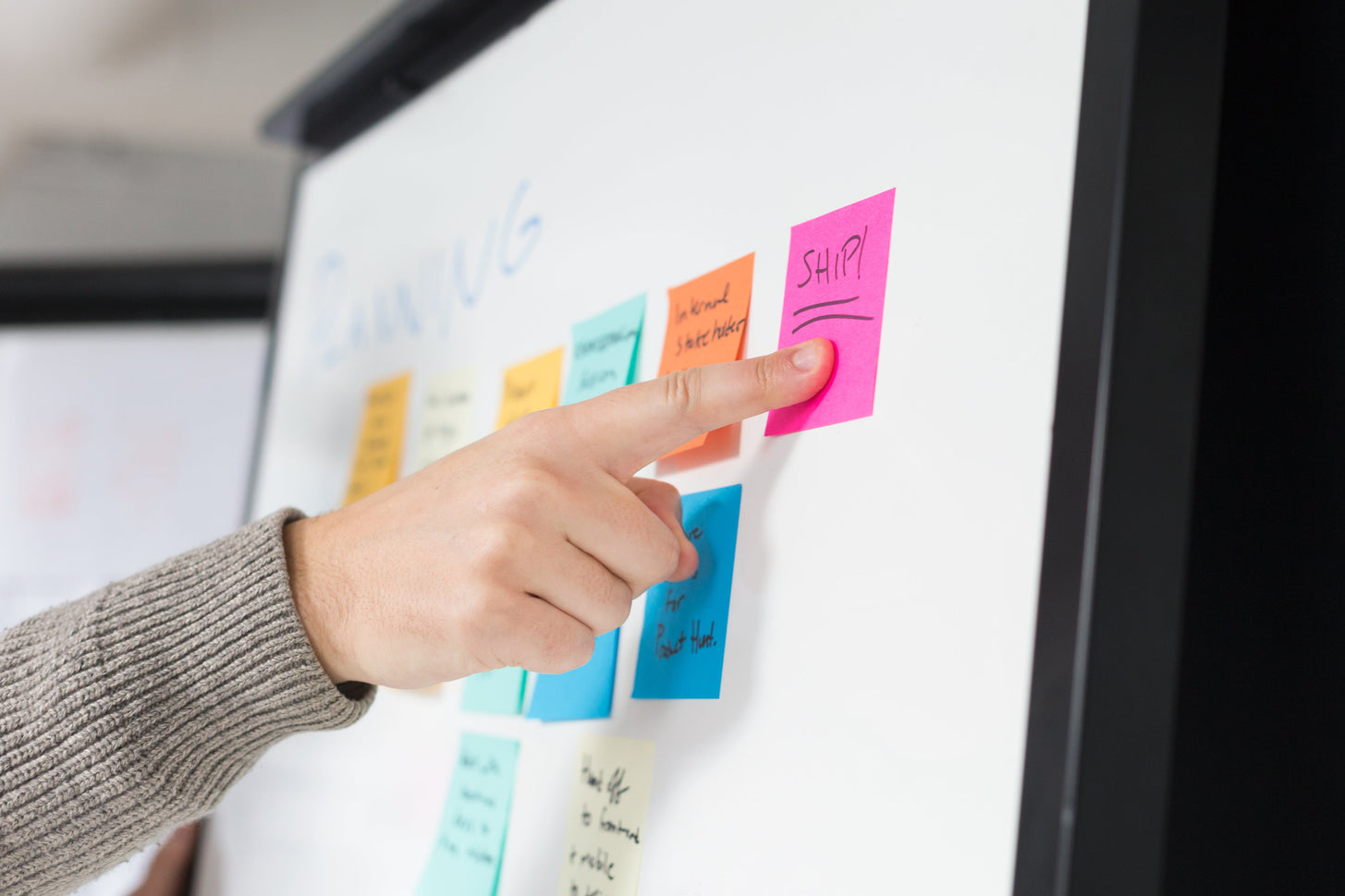 An image of a finger pointing at a sticky note on a whiteboard.