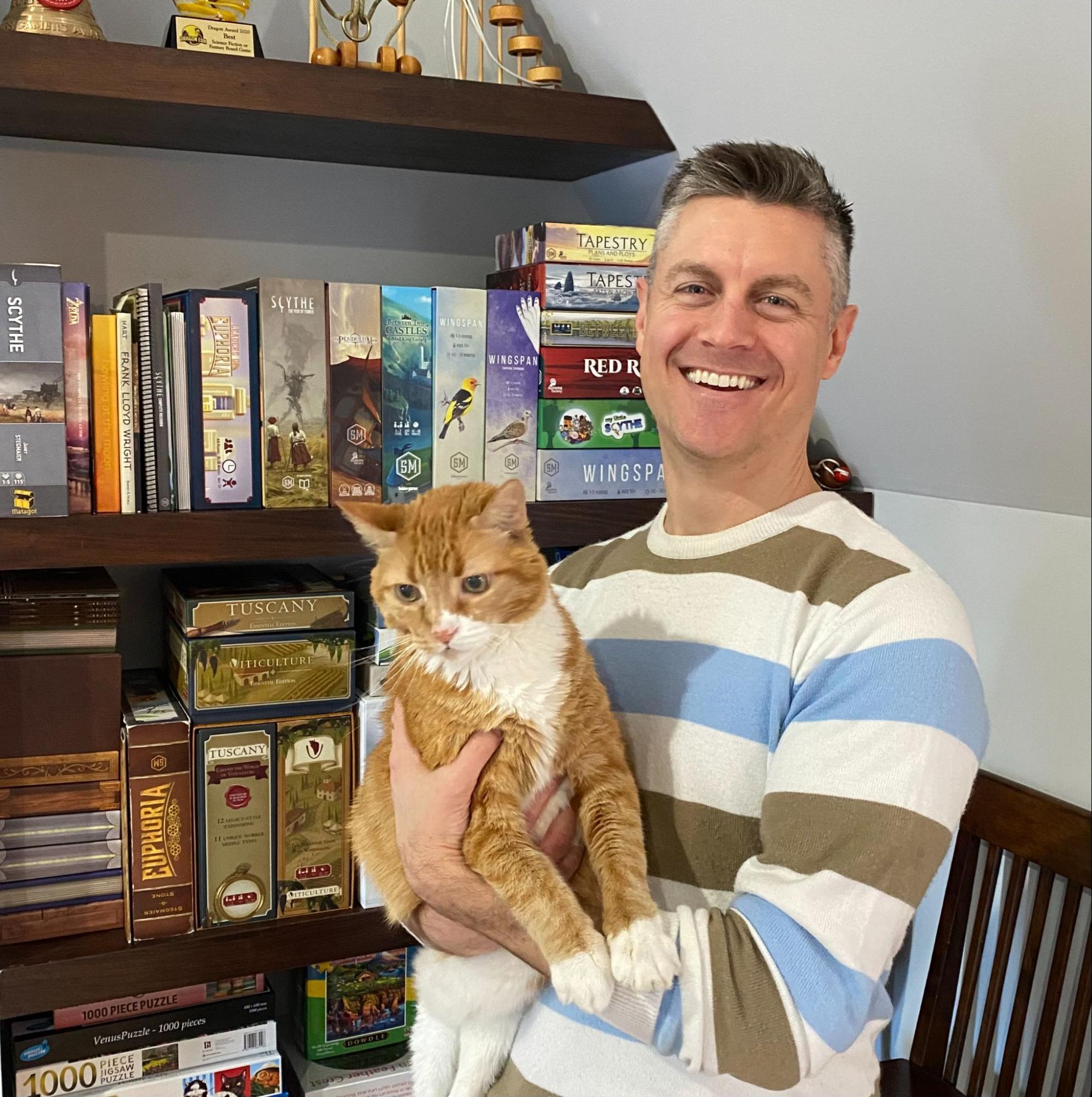 Cofounder of Stonemaier Games, Jamey Stegmaier wearing a striped sweater, holding an orange striped cat standing in a room with a shelf filled with board games. 