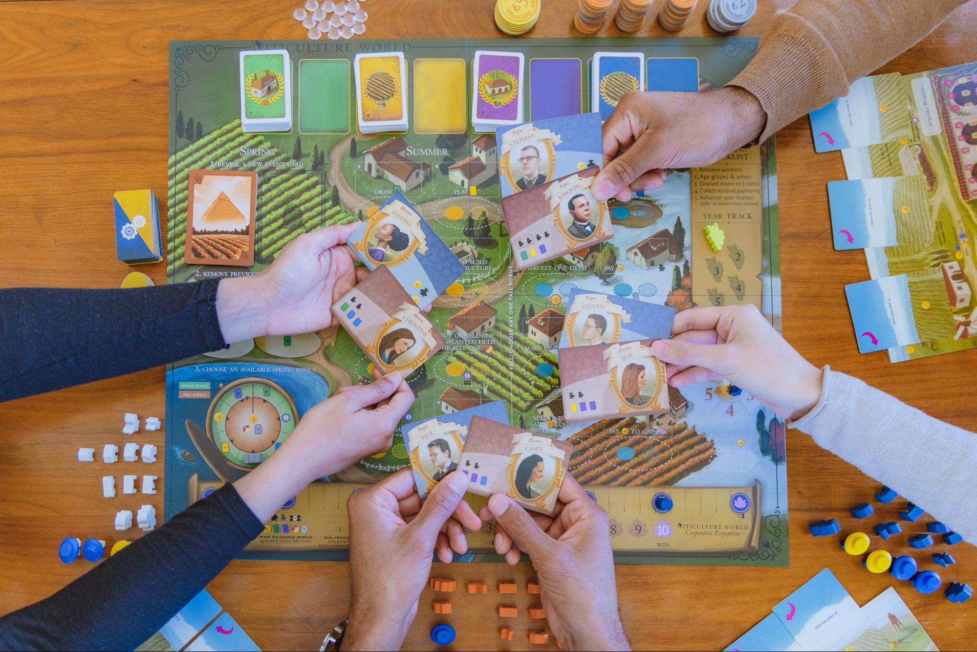 Various hands of board game players holding out cards against the backdrop of the Viticulture board game. 