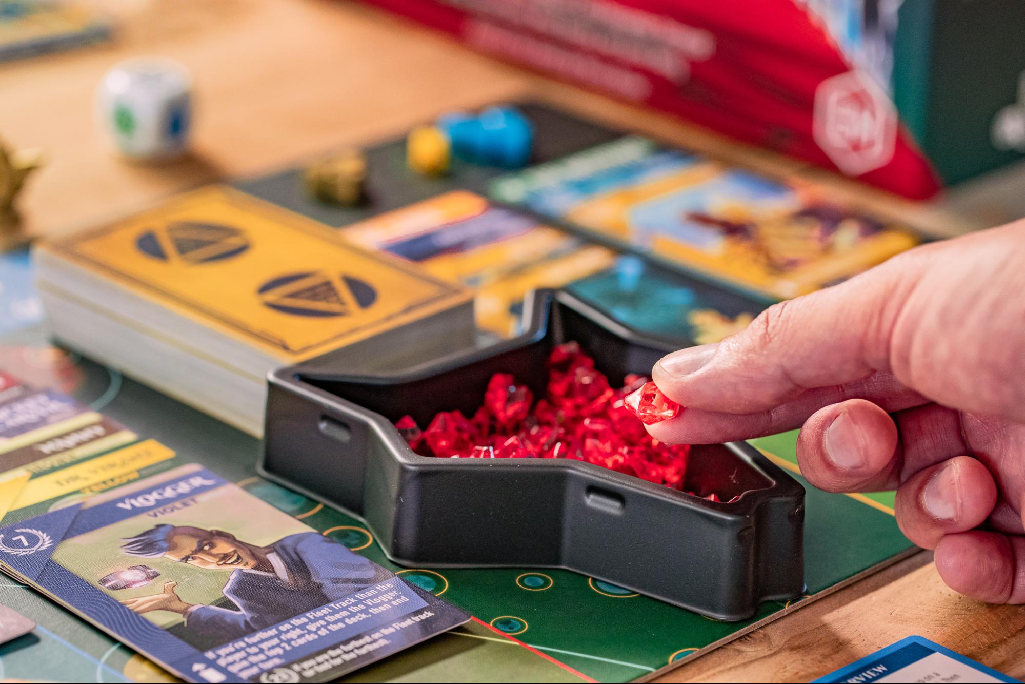 A player holds up pieces of the game Red Raising along with cards and other elements of the board game in the background. 