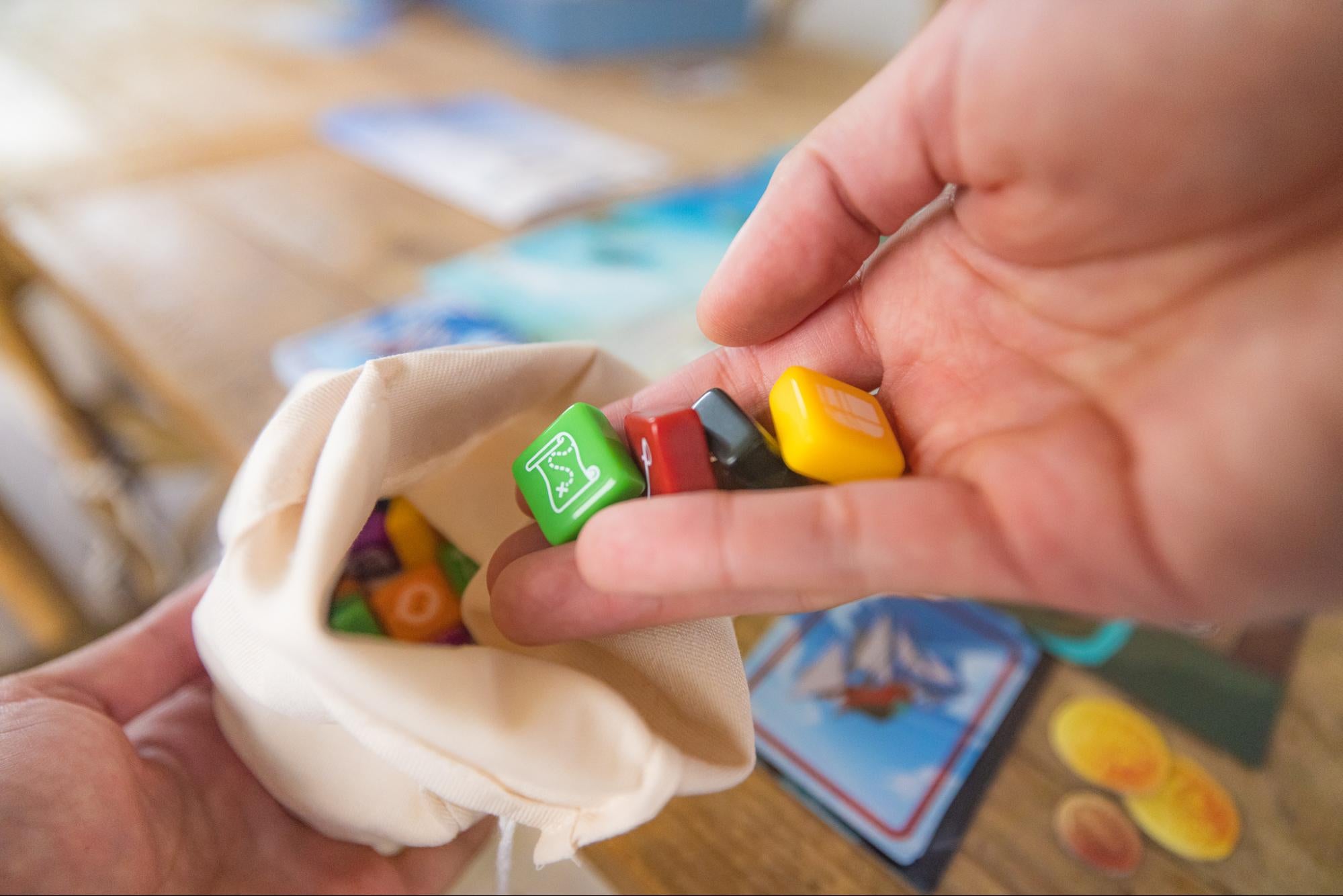 A board game player holds out a fabric bag filled with various game pieces against a table backdrop. 