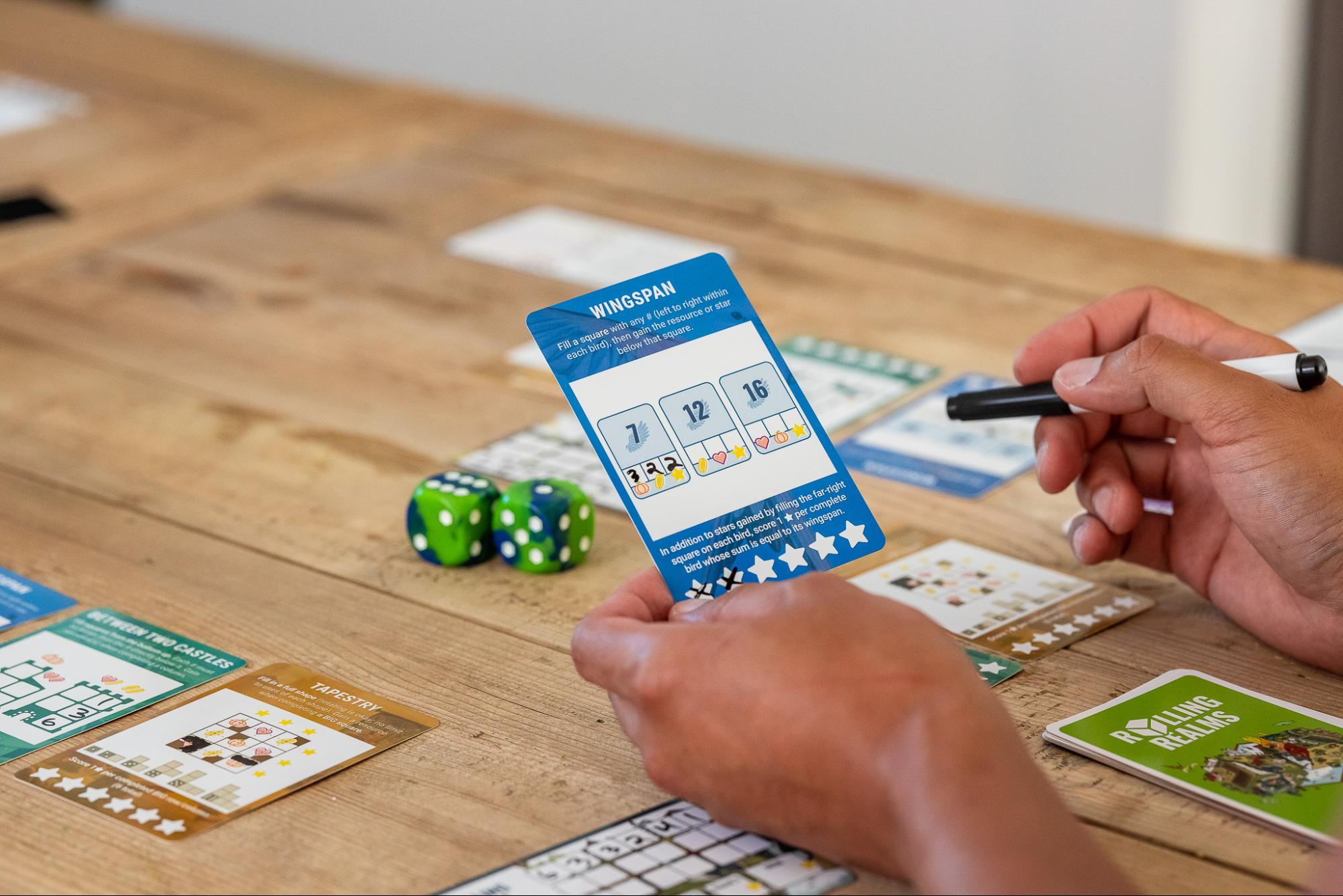 A player’s hands hold up a card from the game Wingspan backdropped by a wooden table with various cards from the game. 