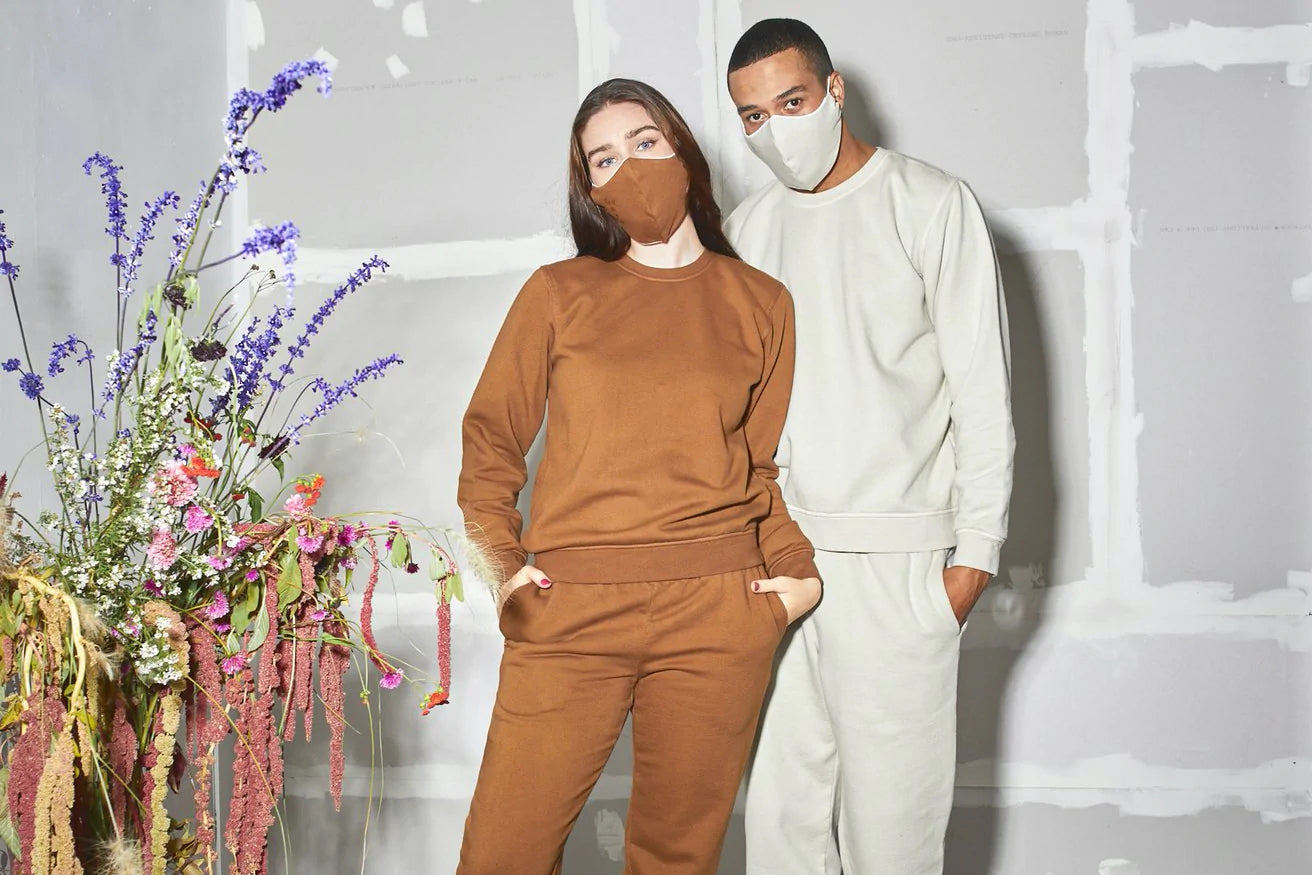 Two people in masks and loungewear pose beside some plants