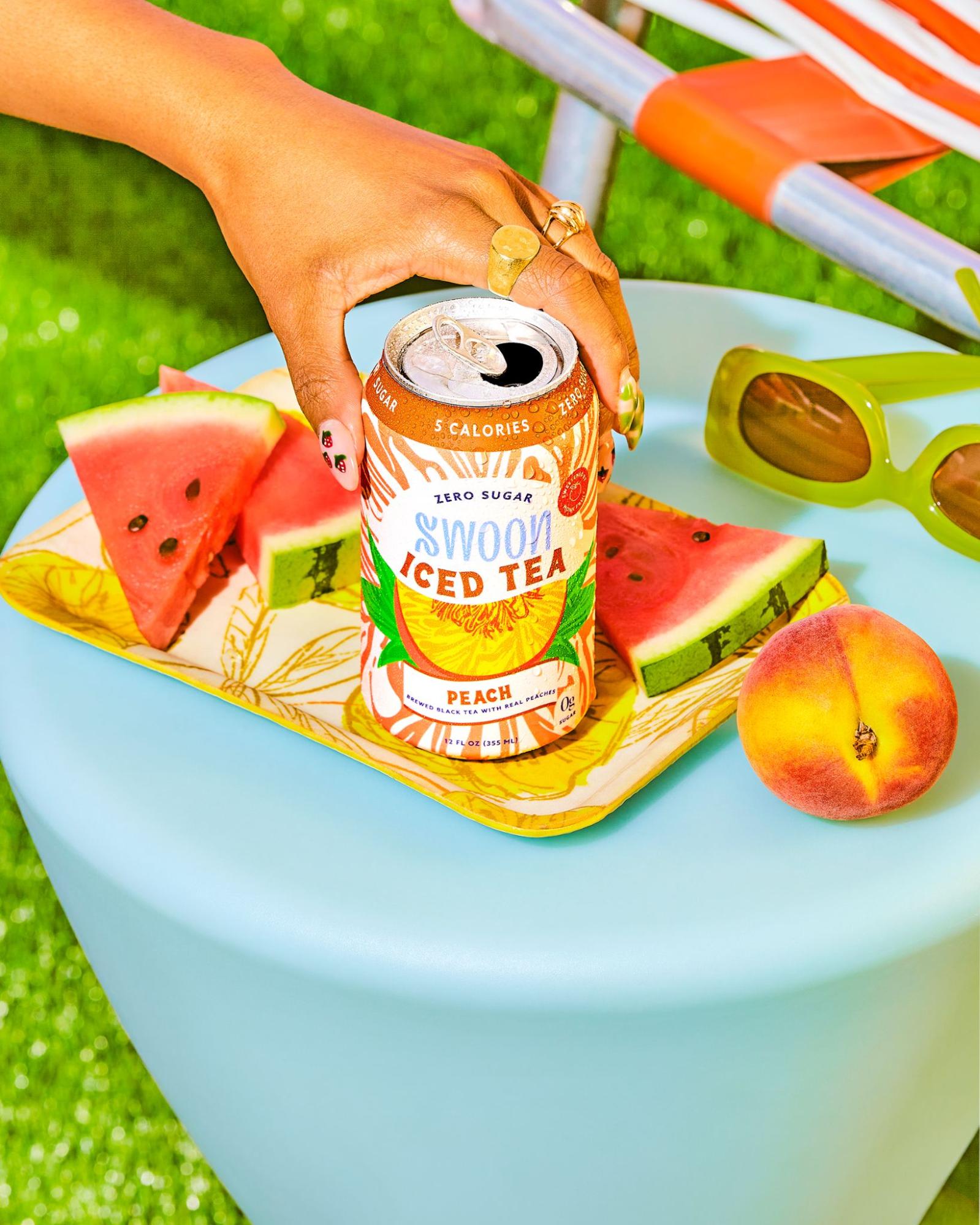 A peach-flavored iced tea by Swoon is placed on a blue table with fruits and sunglasses in the background. 