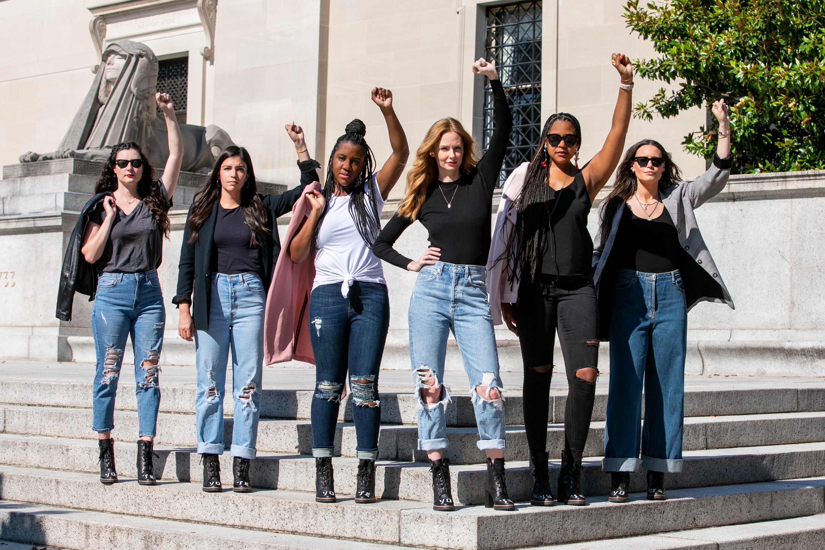 Six women stand on cement steps, all with fists raised in the air