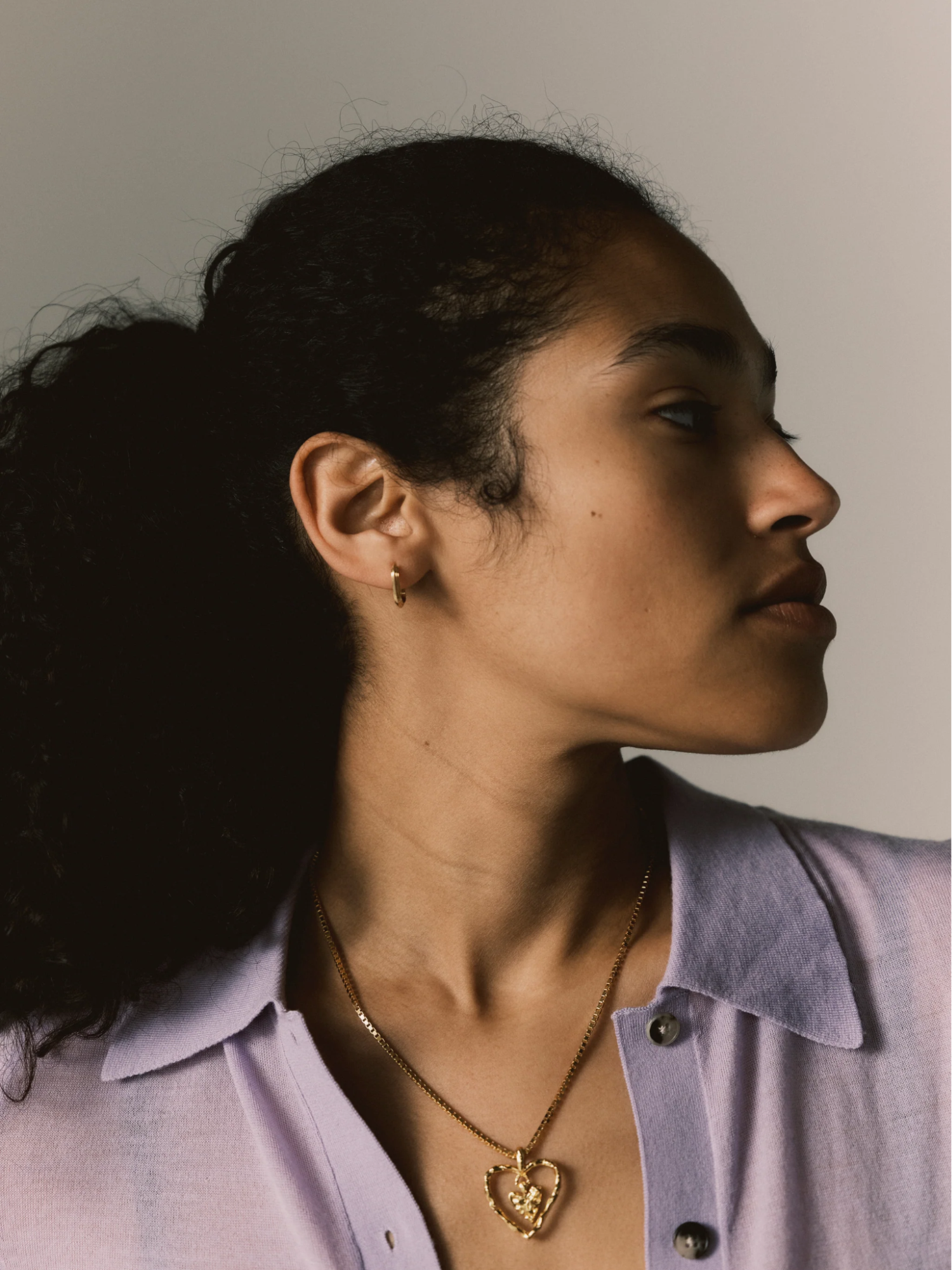 Woman in lavender top wearing a gold heart pendant necklace and small gold hoop earring.