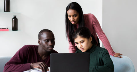 Three people view single laptop computer on desk in modern room