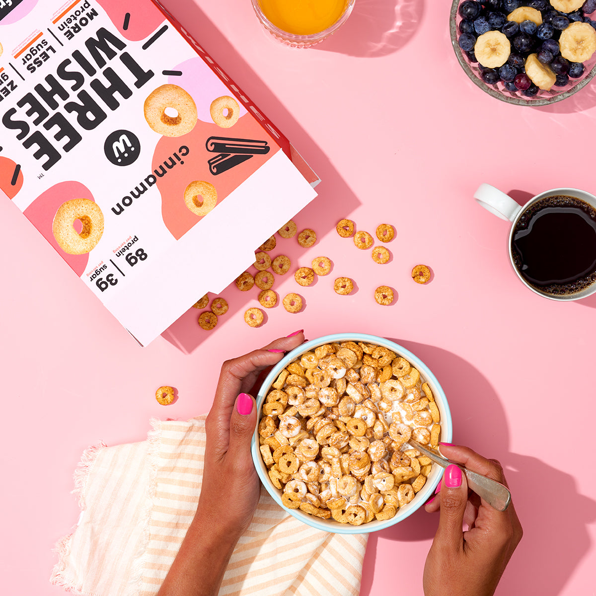 A hand model holding a bowl of cinnamon flavoured cereal against a pink backdrop with coffee, bowl of fruits, and the cereal box in the same frame. 
